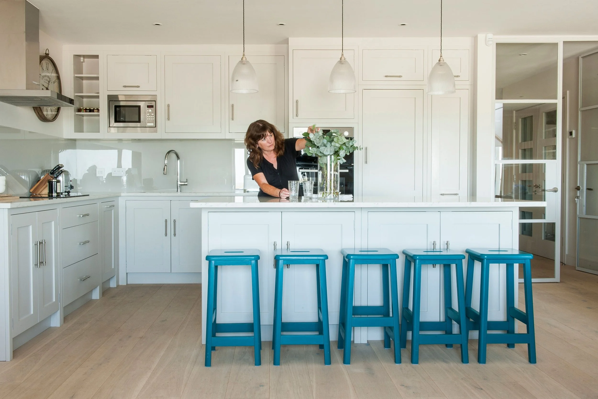 A woman arranging flowers in a kitchen with white cabinets, a kitchen island with five blue stools, and pendant lights hanging from the ceiling.