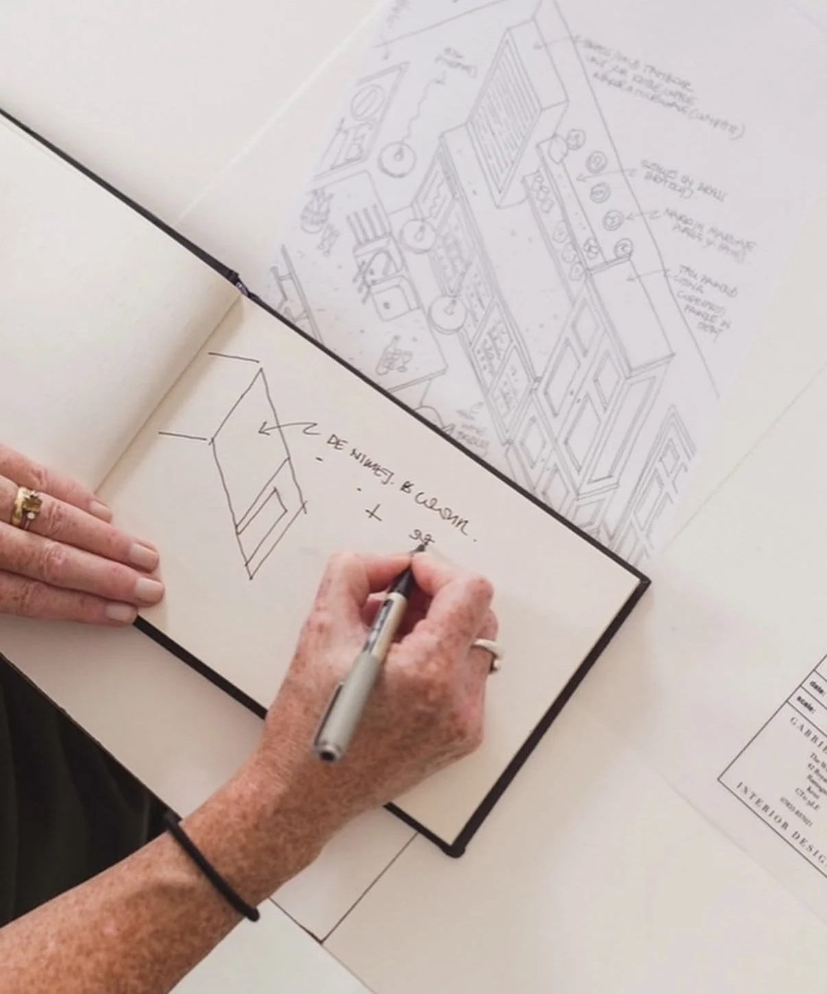 Person drawing a simple sketch of a building plan in a notebook, with architectural blueprints visible on the table.