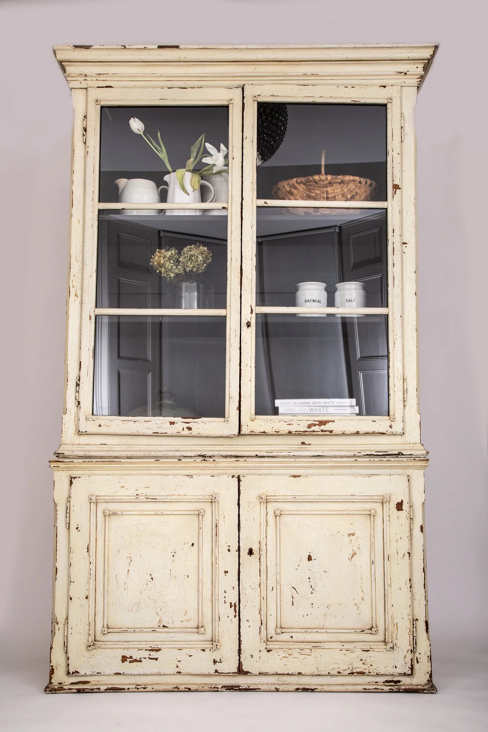 A vintage distressed white wooden cabinet with four glass-paneled doors, containing decorative items such as white ceramic vases, a basket, books, and a jar labeled 'Oatmeal' and 'Salt'.