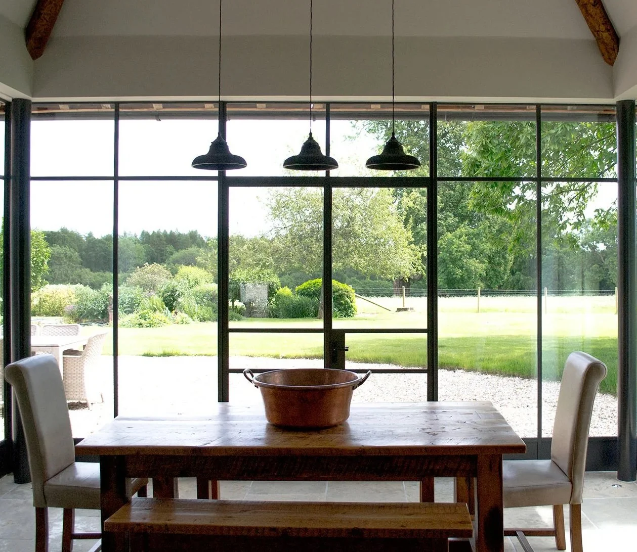 Interior view of a dining room with a wooden table and two white chairs, overlooking a lush green garden through large glass windows, with three hanging ceiling lights.