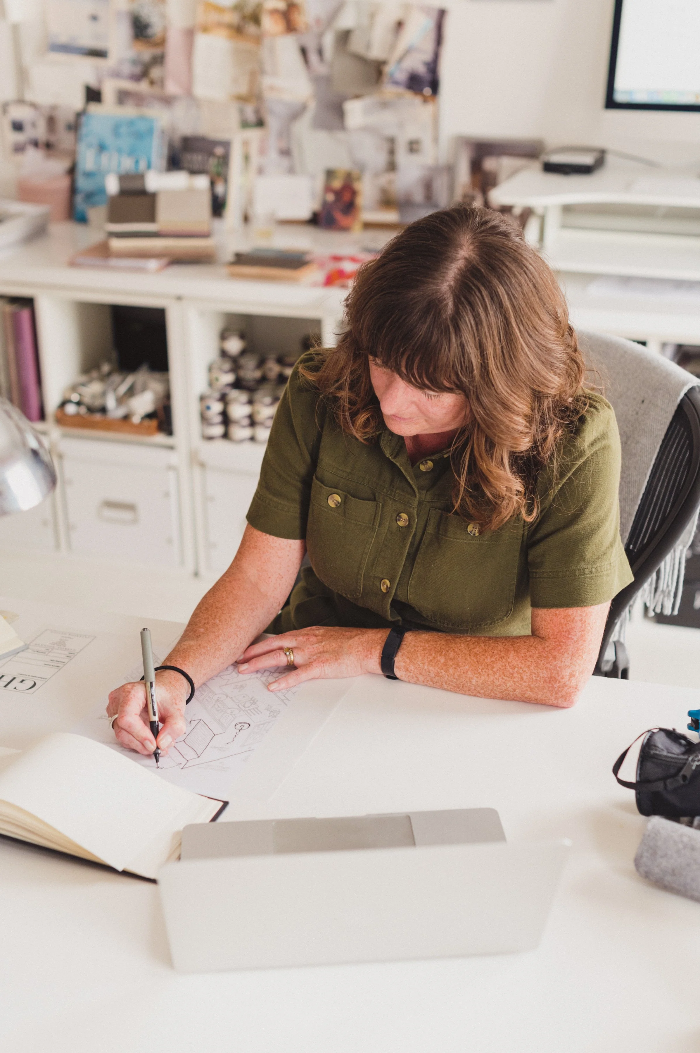 A woman with brown hair and freckles, wearing an olive green shirt, is sitting at a white desk, drawing sketches on paper with a black pen.