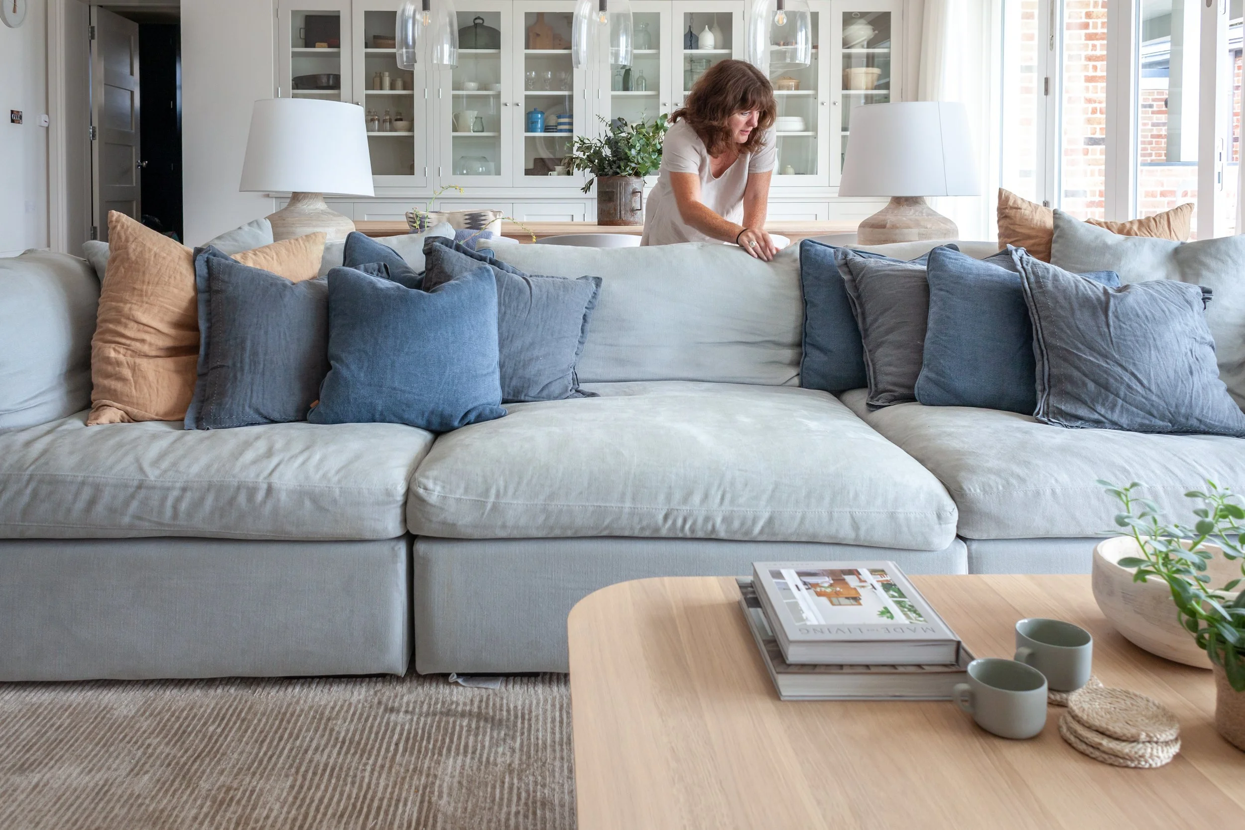 A woman arranging a large, cream-colored sofa with blue and beige pillows in a bright living room with white built-in shelves, glass cabinets, and a wooden coffee table with magazines, mugs, and ceramics.