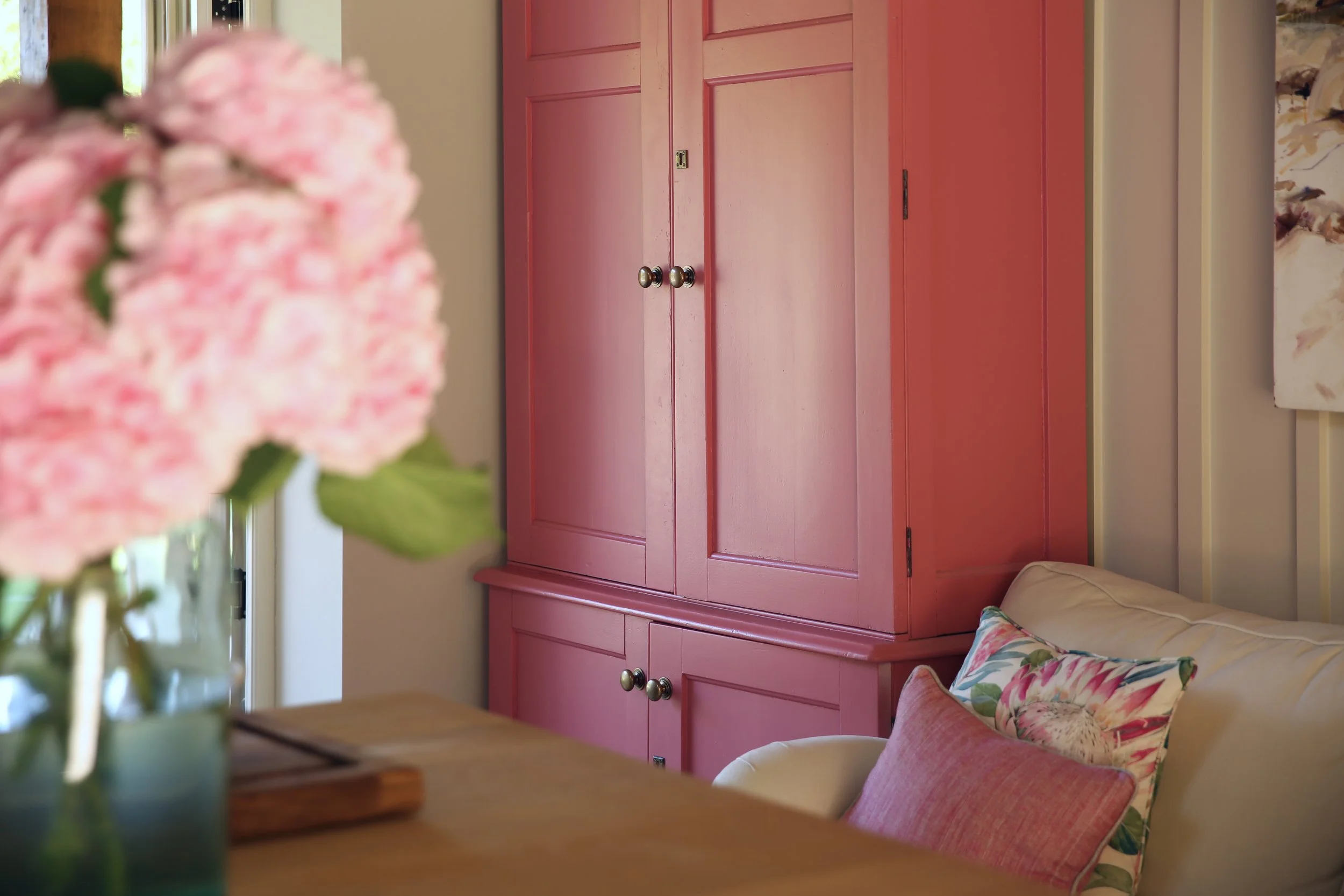 A pink wooden cabinet with rounded brass knobs in a home interior. Part of a floral-patterned sofa and a cushion are visible in the foreground.