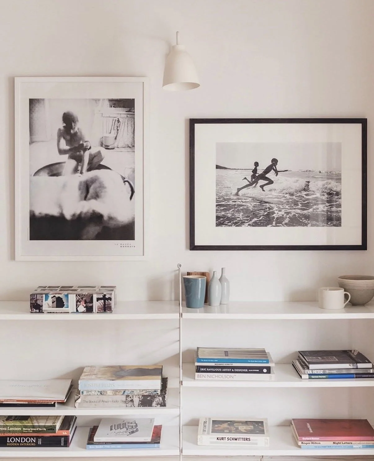 A white bookshelf with books and decorative items, two framed black and white photographs of children at the beach on the wall above, and a hanging white lamp.