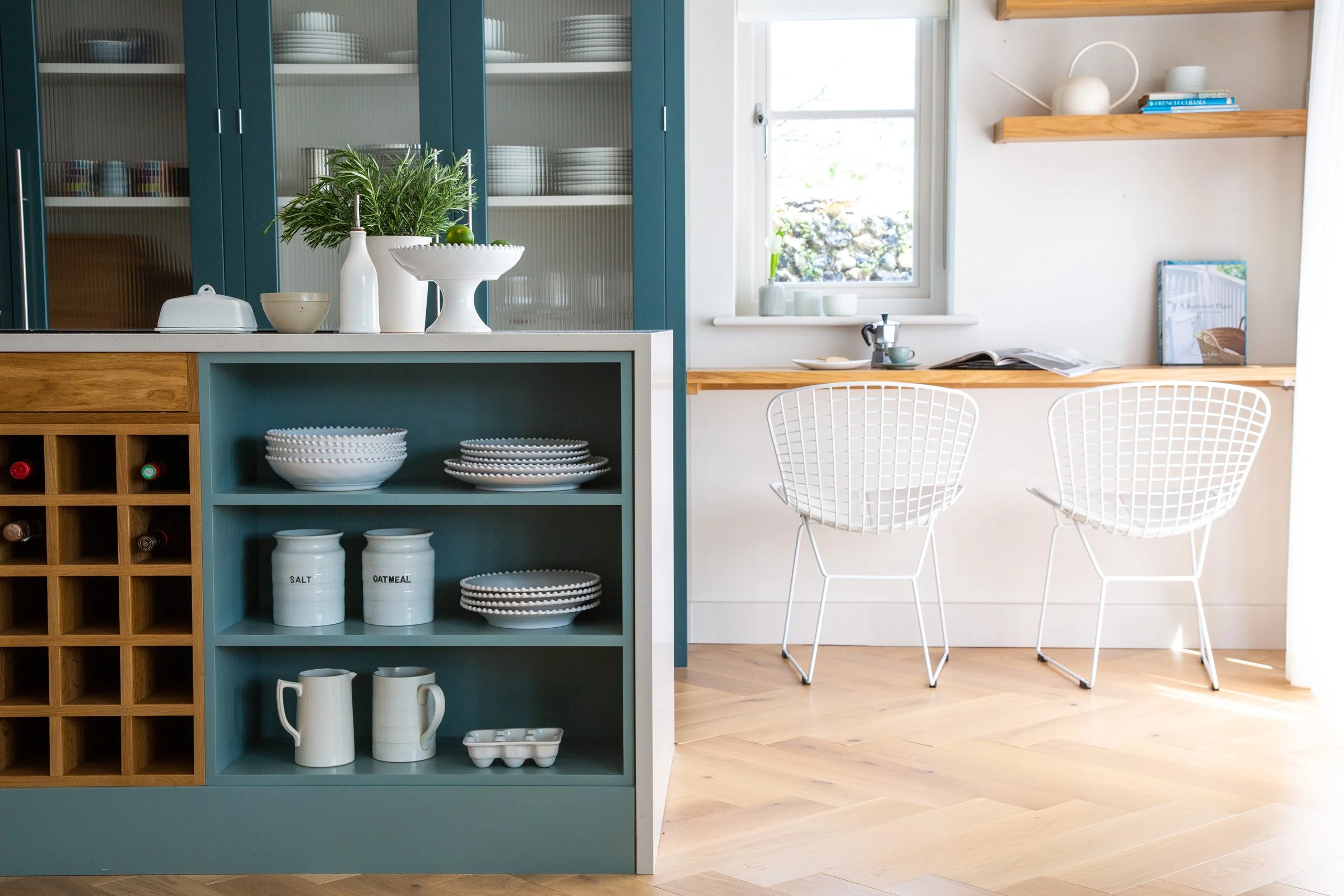 A bright, modern kitchen with a blue open shelving unit displaying white dishes, bowls, and vases. There is a wine rack on the left below the shelves. In the background, cream-colored walls feature a wooden shelf with books and decorative items, and a window with sunlight streaming in. Two white wire chairs are positioned in front of a wooden work surface.