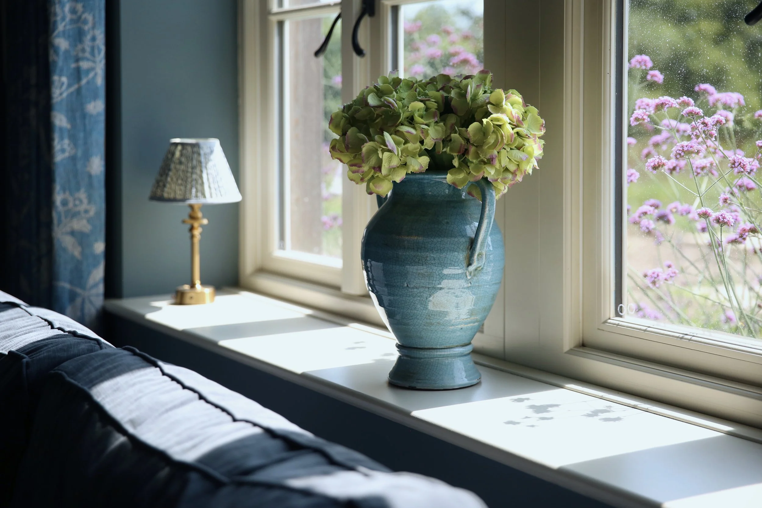 A blue ceramic vase with pink and green hydrangea flowers on a windowsill. Outside the window, pink flowers and greenery are visible, and raindrops are on the glass. A small table lamp with a patterned shade is next to the vase.