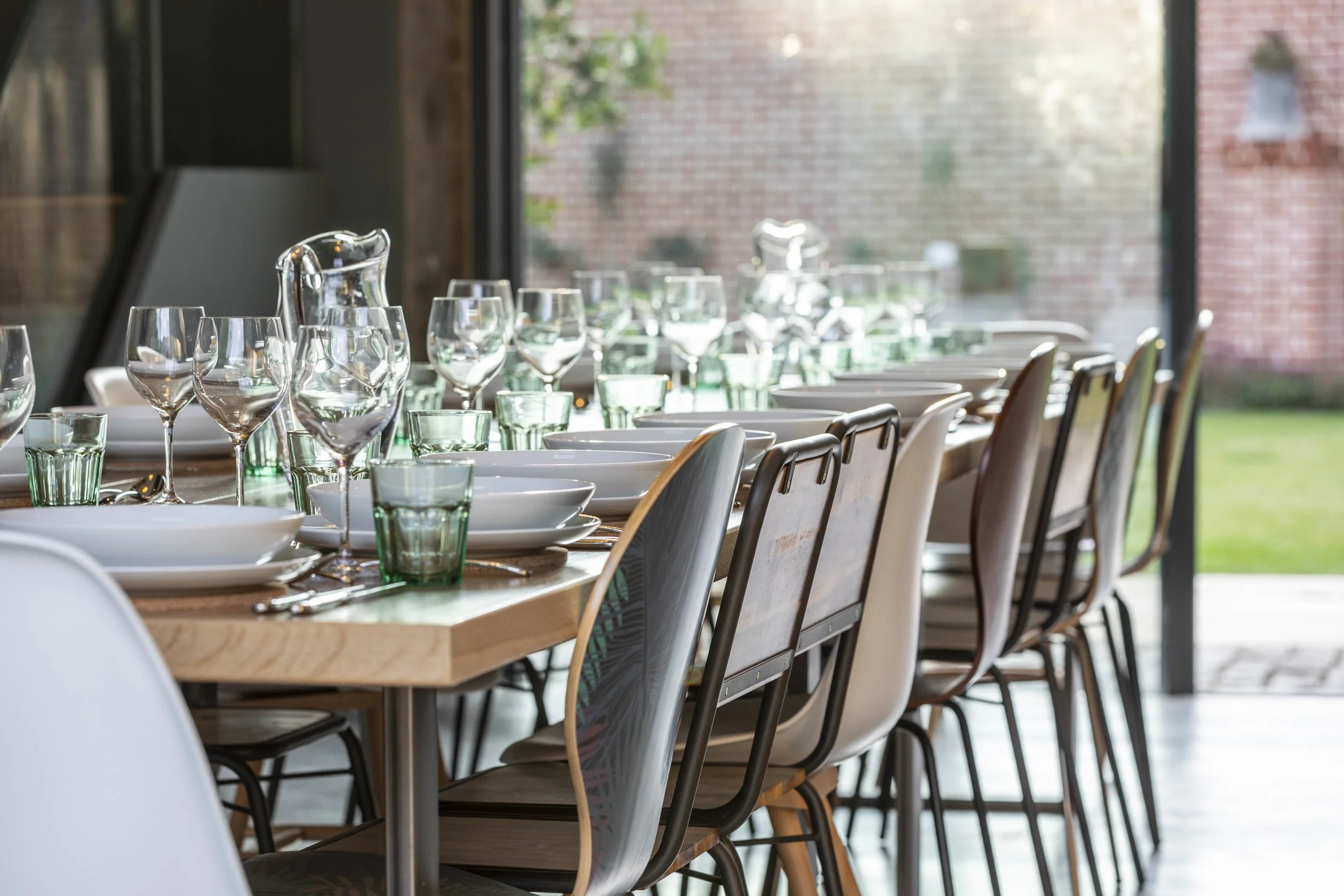 A long dining table set with wine glasses, water glasses, white plates, and silverware, arranged for a meal in a room with large windows showing an outdoor brick wall and greenery.