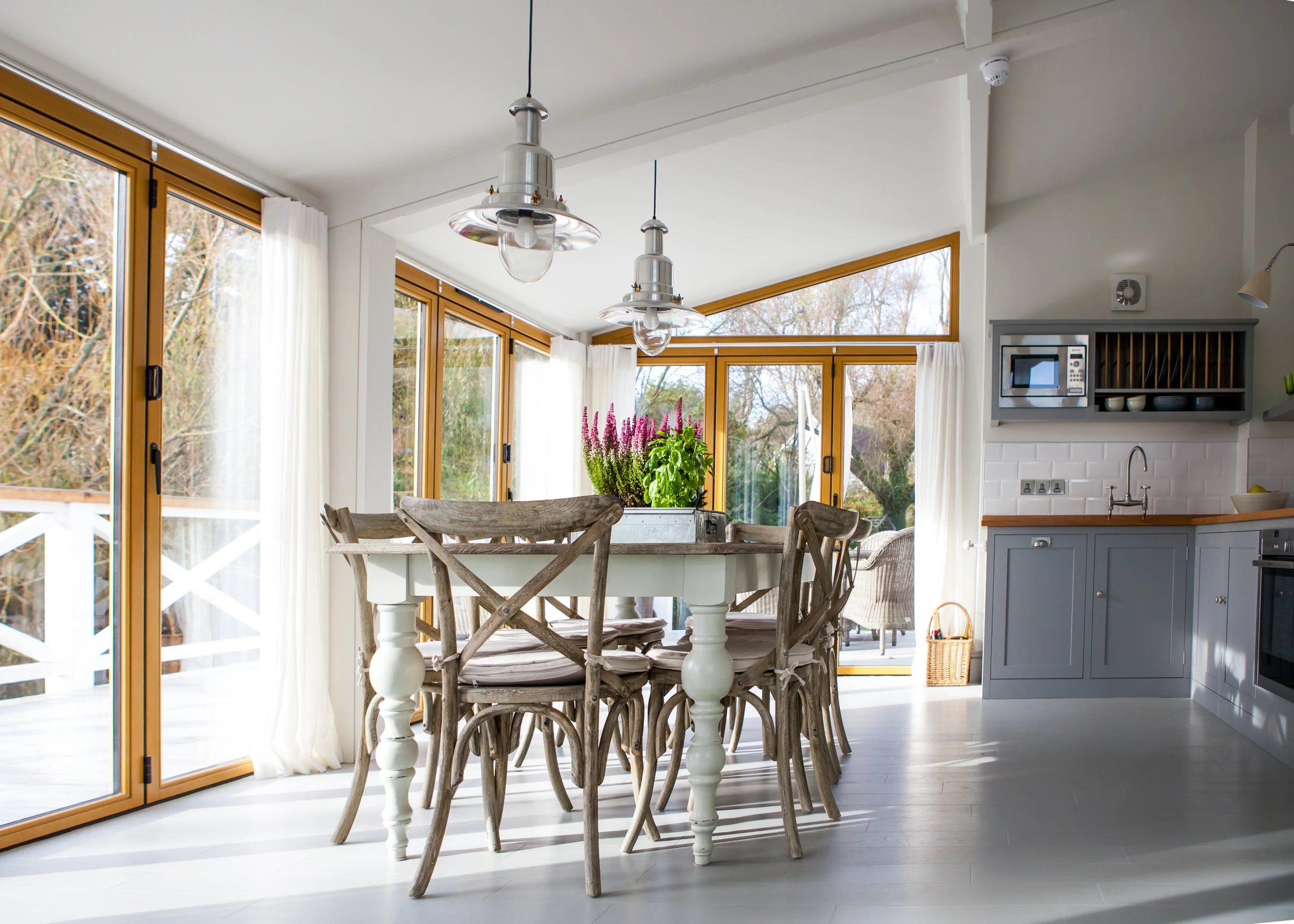 A bright dining area with a white table and rustic wooden chairs, large windows with white curtains, and modern light fixtures hanging from the ceiling. There are trees visible outside through the windows.