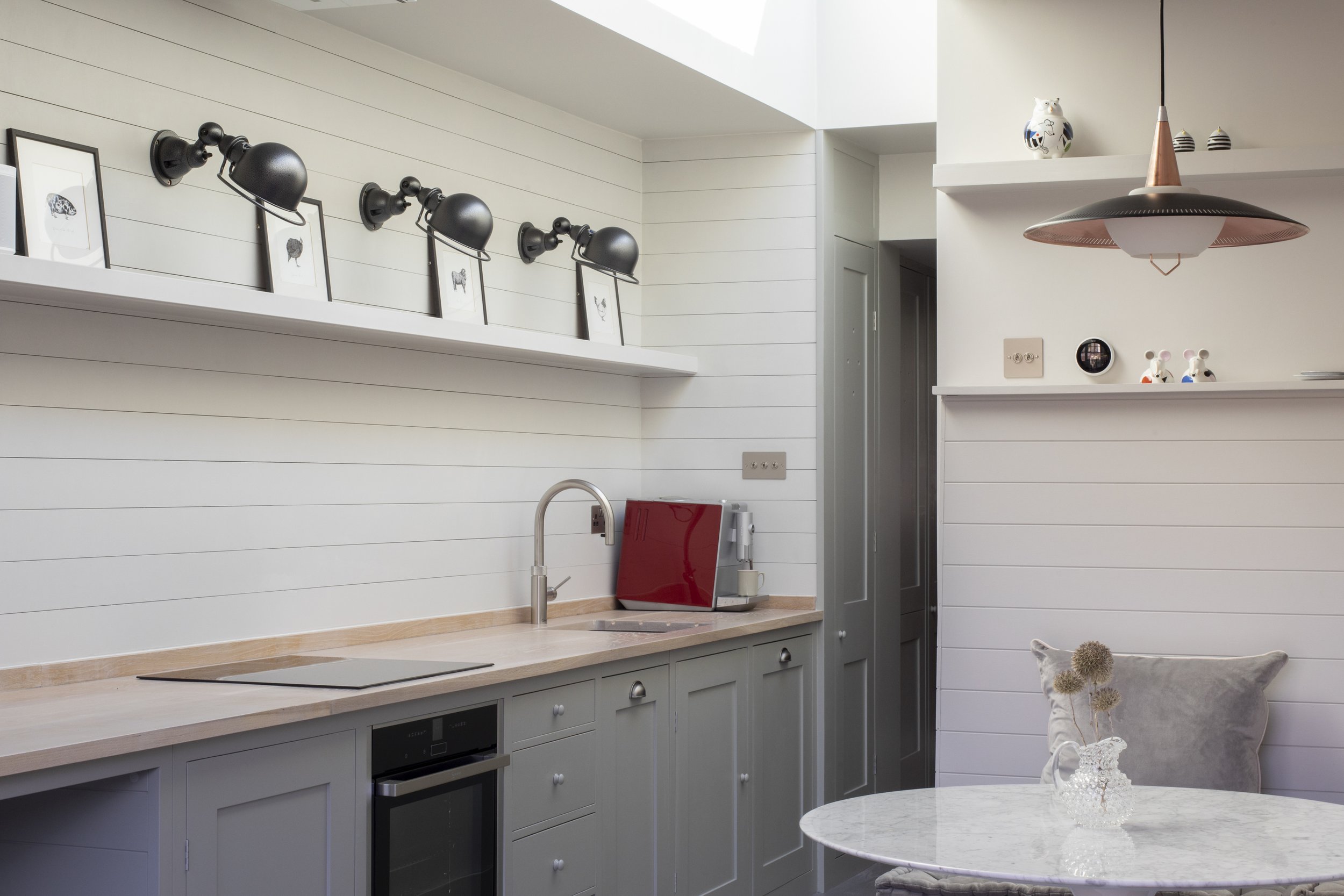 A modern kitchen with white shiplap walls, light wood countertops, and gray cabinets. Black wall-mounted lamps, framed artwork of animals, a red toaster, and decorative items including mice figurines and a ceramic owl are on the shelves. A silver faucet, a round table with a white marble top, and a bowl of dried flowers are also visible.