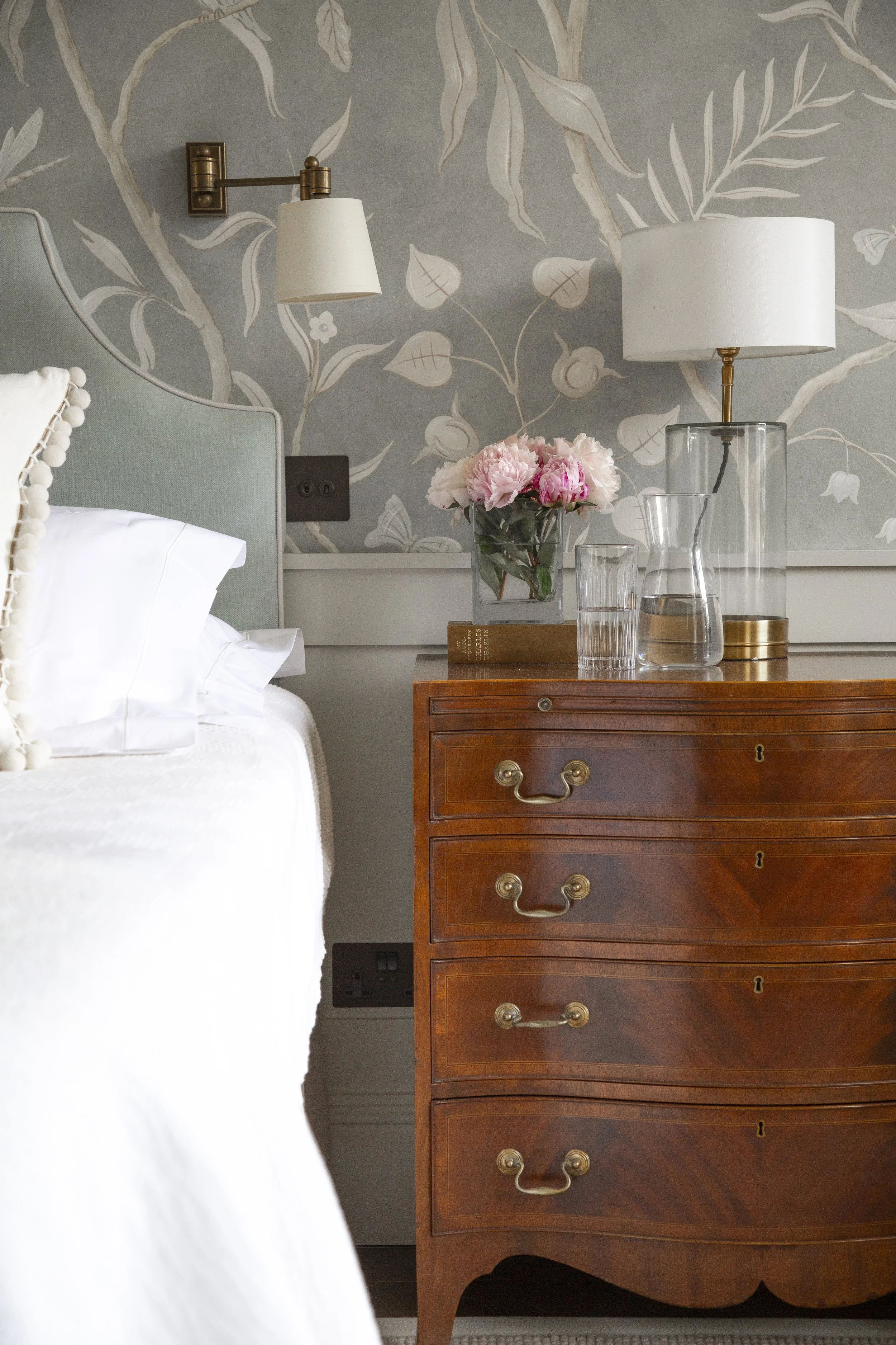 A bedroom nightstand with a vase of pink peonies, a glass pitcher, a glass, a table lamp, and a floral wallpaper with a grey background.
