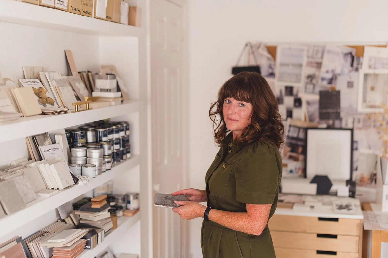 A woman with shoulder-length brown hair holding a sample tile stands in a design studio, surrounded by shelves of paint swatches and samples on the wall.