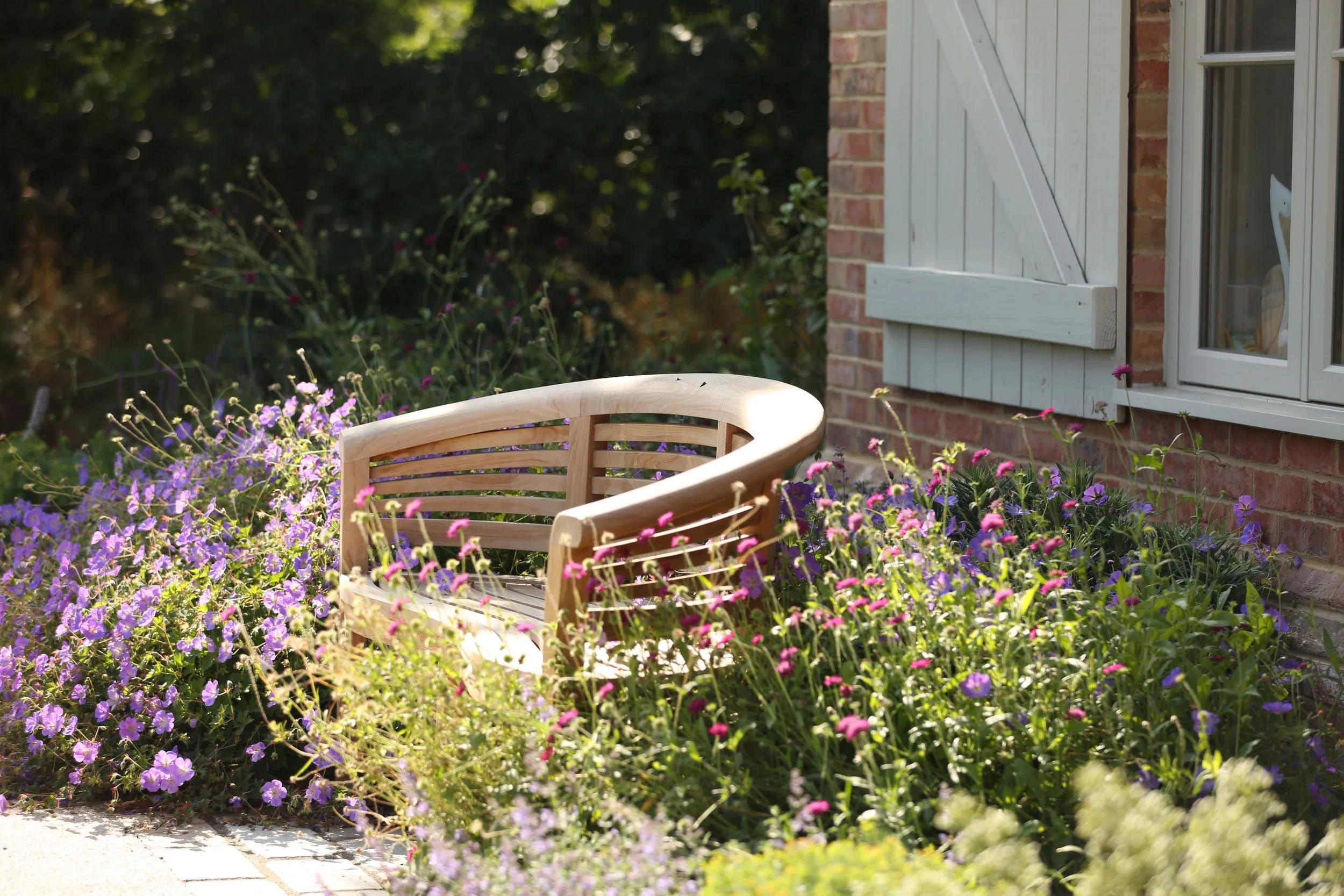 A garden with purple and pink flowers in front of a brick house with white window shutters.