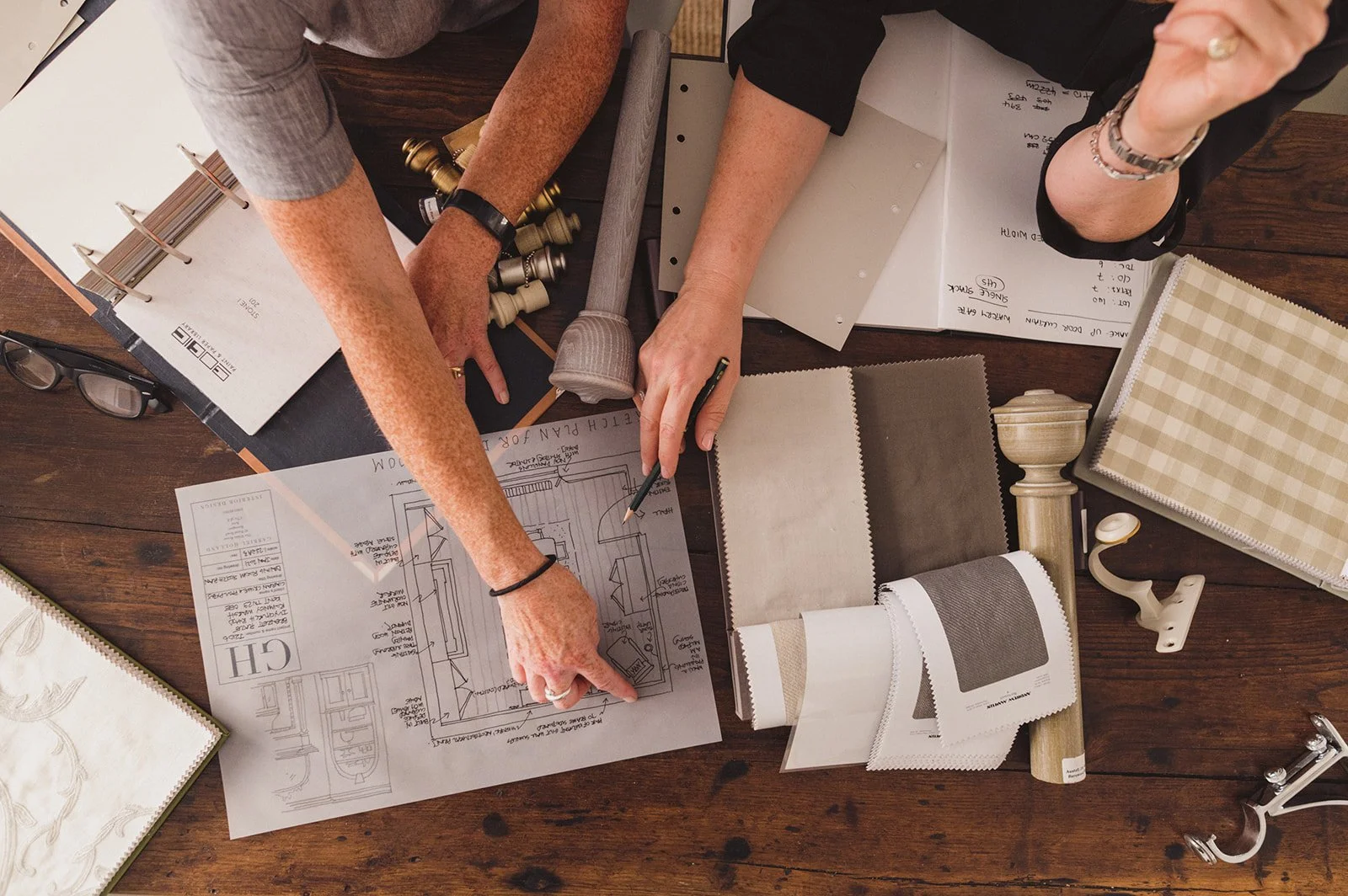 Two individuals collaborating on an interior design or home renovation project, examining architectural plans and fabric samples on a wooden table.