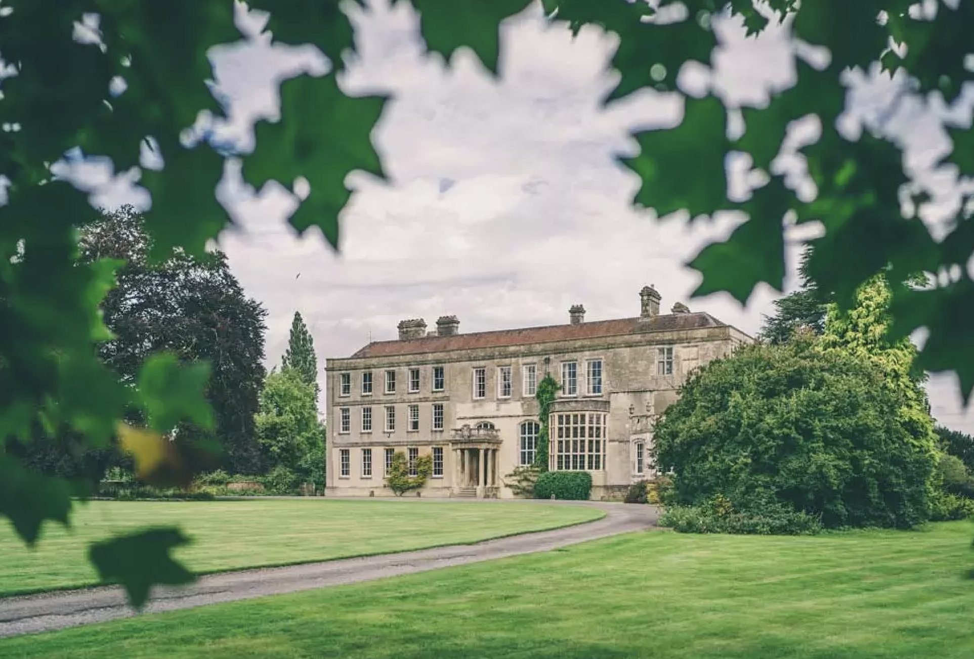 A large, elegant mansion with multiple chimneys and large windows is framed by lush green trees and bushes. The mansion is situated on a well-maintained lawn with a curved pathway in the foreground, under a partly cloudy sky.