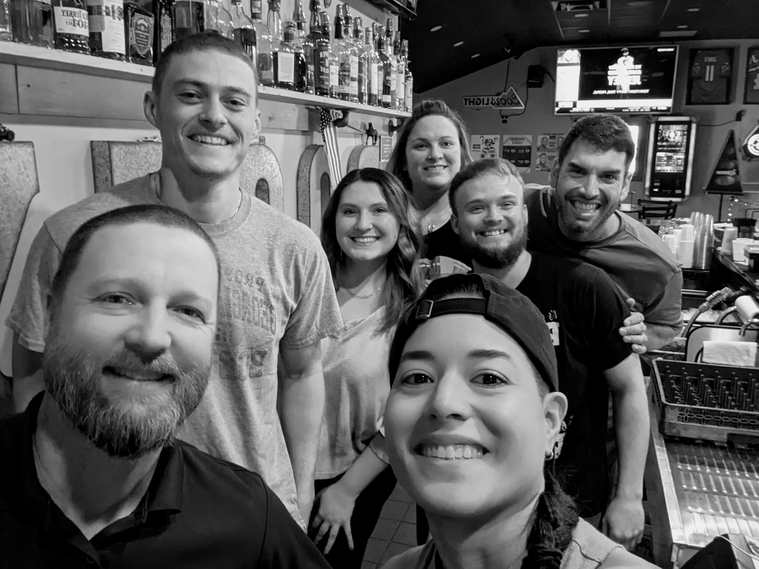 Group of six friends smiling inside a bar or restaurant, with bottles of alcohol on the shelves behind them.