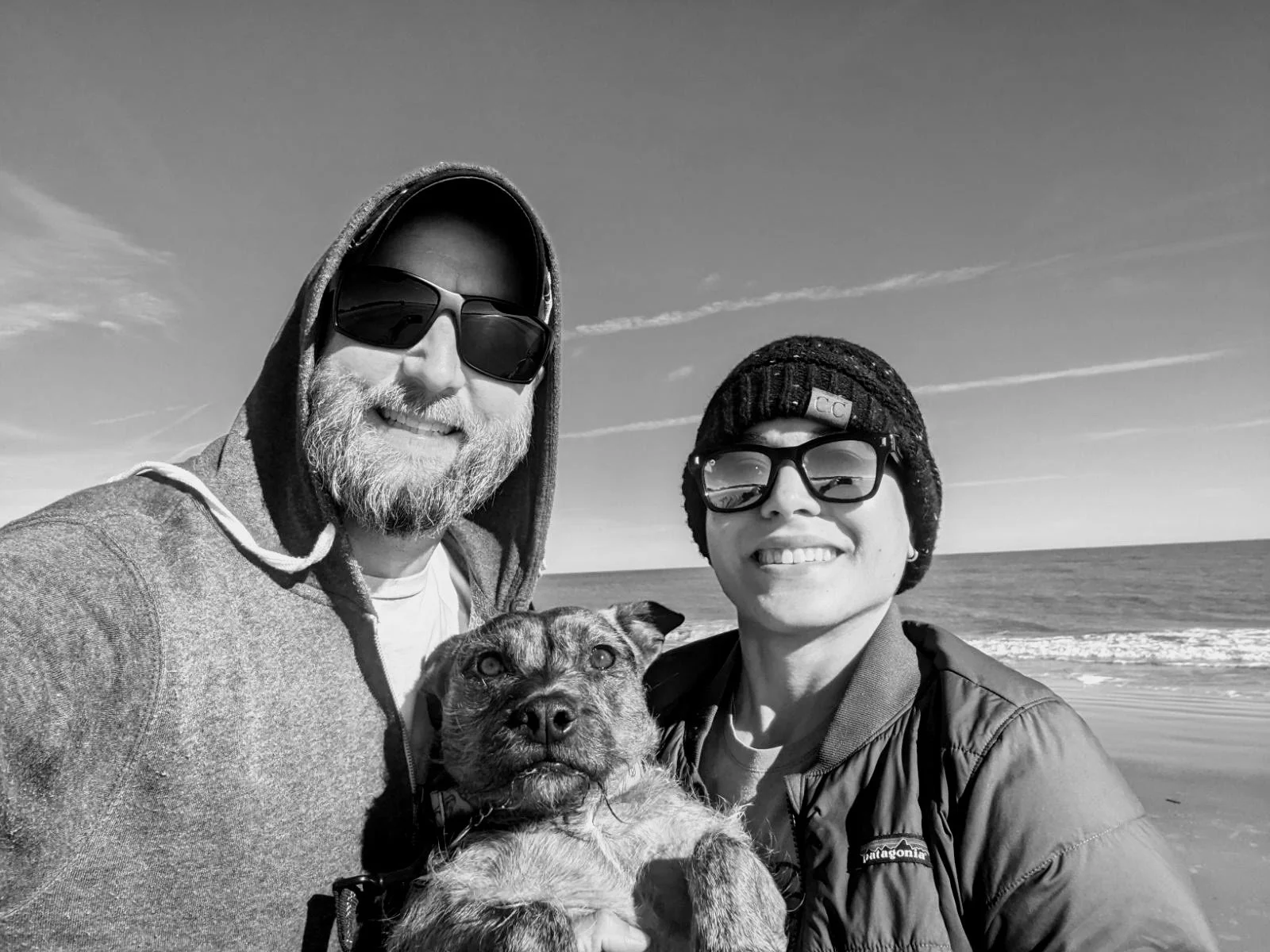 A black and white photo of two men and a dog at the beach, smiling and wearing sunglasses, with ocean and sky in the background.