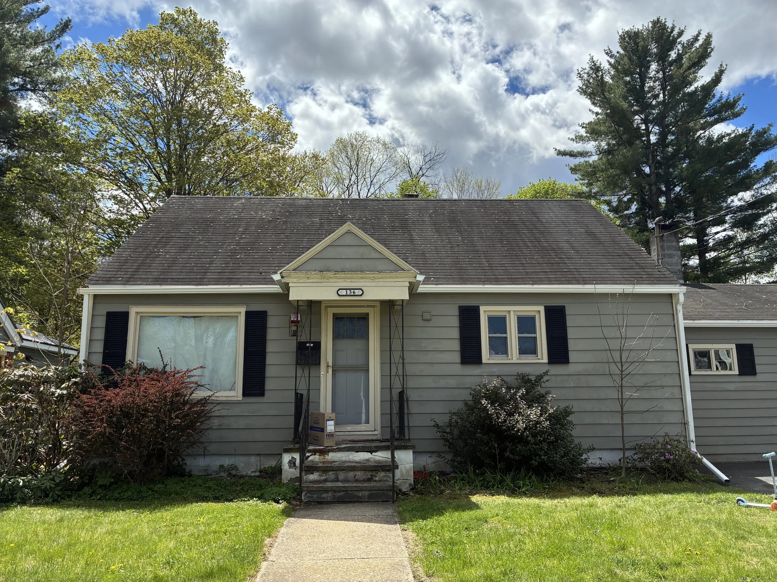 A house with light-colored siding, black shutters, and a gable roof. The house number 136 is displayed above the front door. There are trees surrounding the house, and the sky is partly cloudy with blue patches.