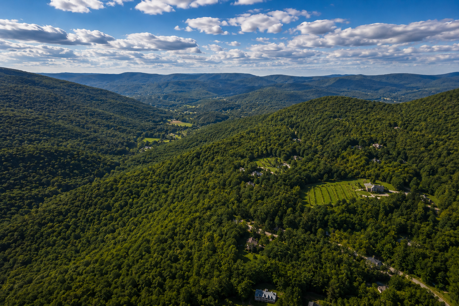 Aerial view of lush green rolling hills with scattered houses, trees, and small farms under a partly cloudy sky.