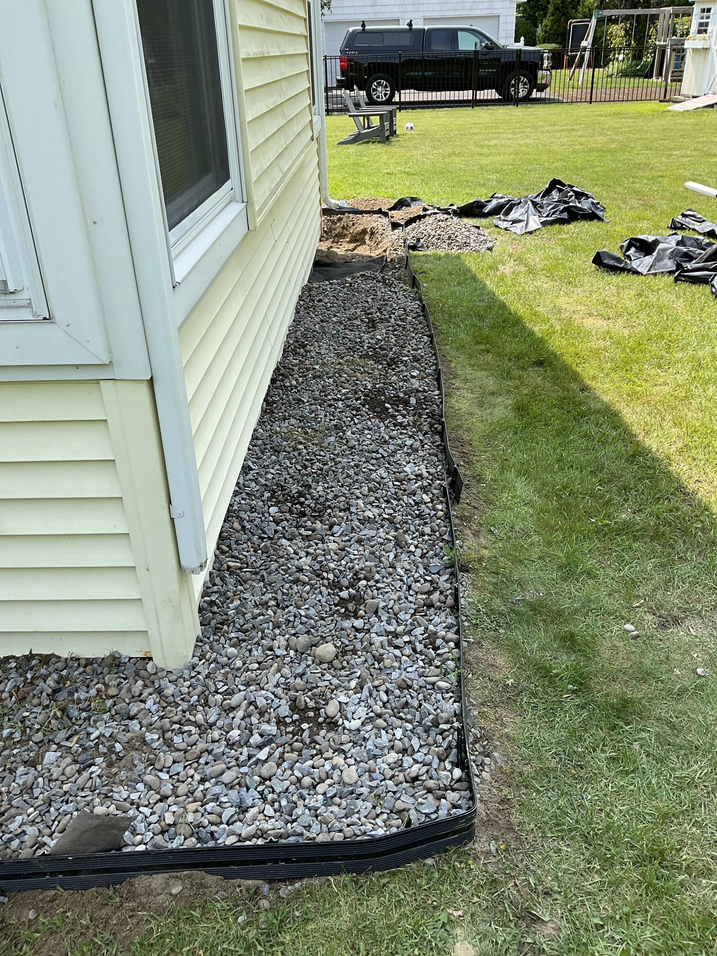 Side yard with gravel and landscaping border alongside a house with yellow siding. In the background, a black truck parked behind a fence, with a soccer ball and garden equipment visible.