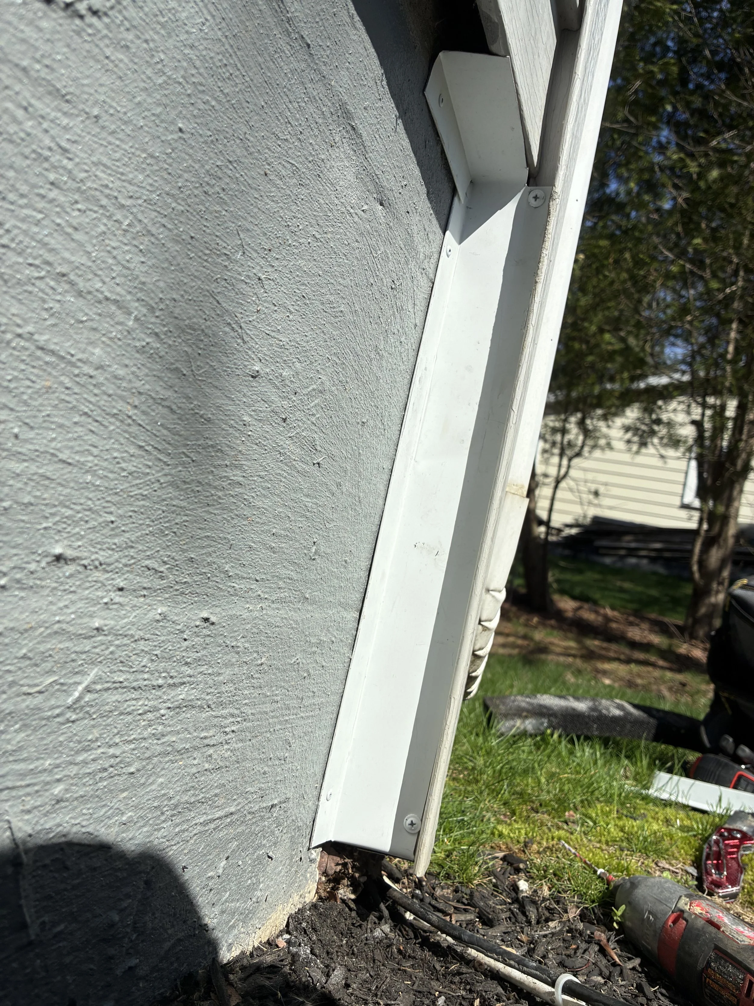 Close-up of a gray textured wall with a white metal box attached, partially buried under soil and debris, with gardening tools and grass in the background.