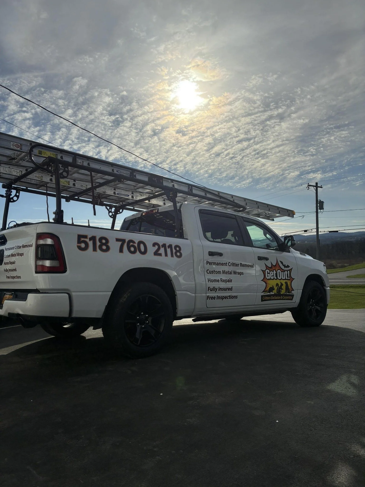White pickup truck with ladder rack, advertisements for critter removal and home repairs, parked on asphalt, under a partly cloudy sky with the sun visible.