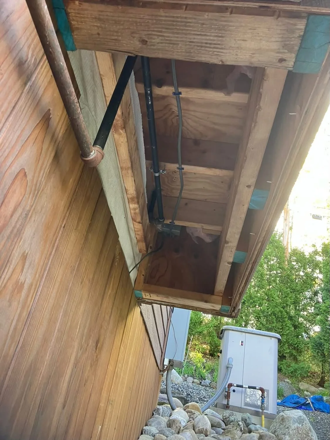 View of an outdoor wooden deck with plumbing and electrical wiring installed under the overhang of a house, with a hot tub and trees visible in the background.