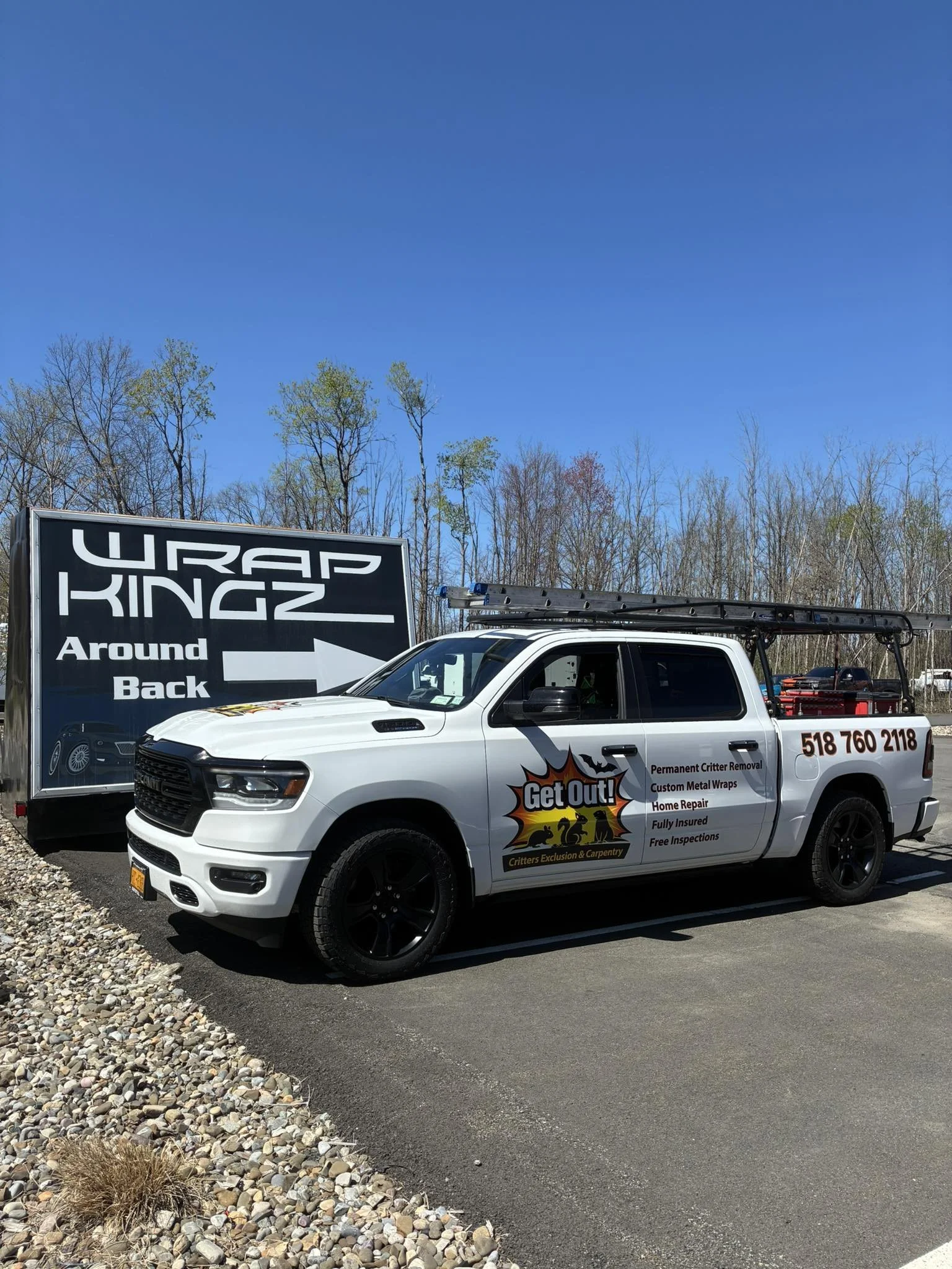 A white pickup truck with black wheels and a ladder on its bed, parked next to a large black sign that reads 'URAP KINGZ Around Back.' The truck has a logo promoting critter removal and pest control services, along with a phone number, and additional services listed such as home repair and free inspections. In the background, there are trees with some green leaves and a clear blue sky.