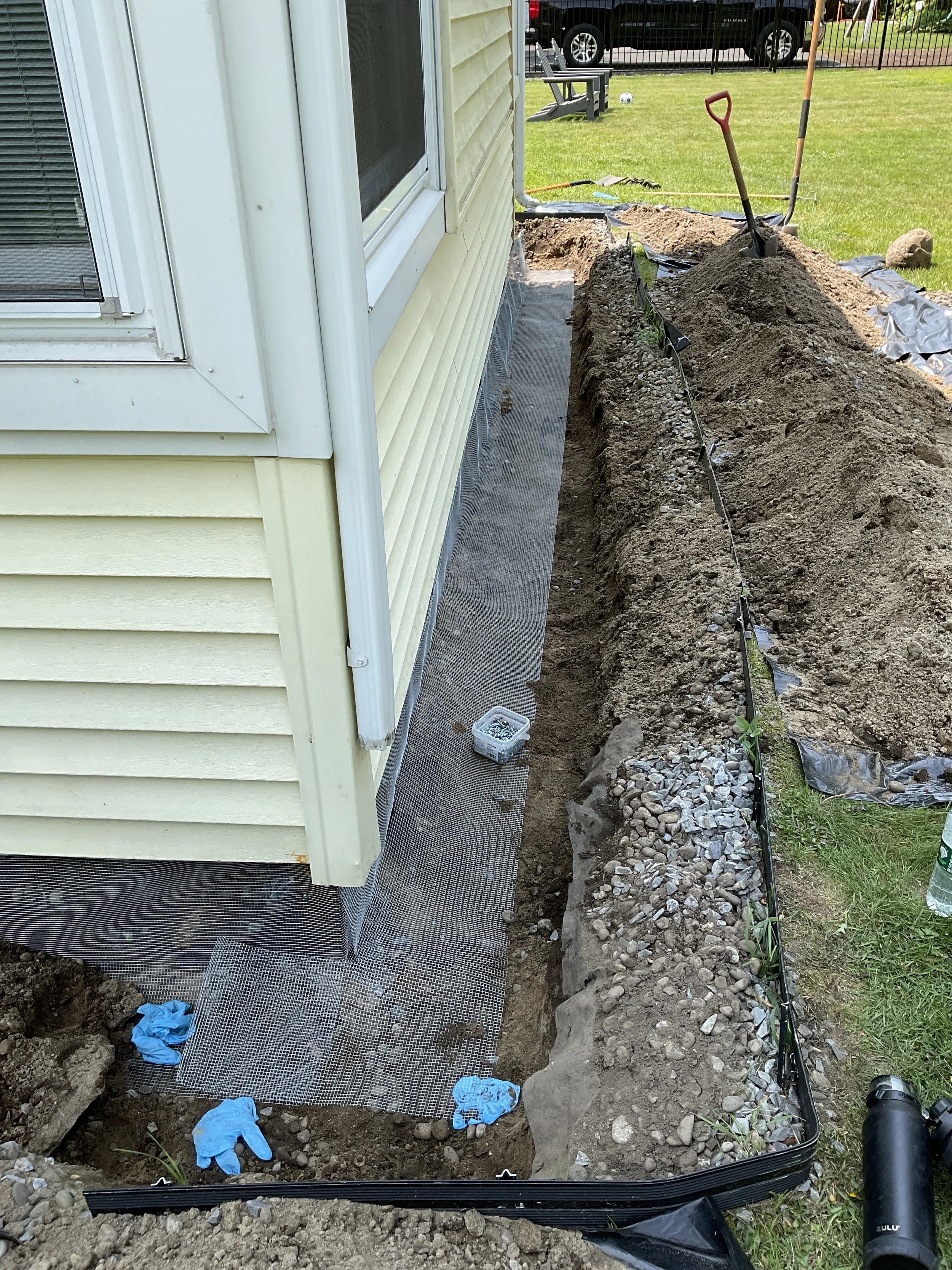 Construction site with trench dug along the side of a house, pants and tools inside the trench,2 shovels standing in the middle, and gravel deposited along the edge.