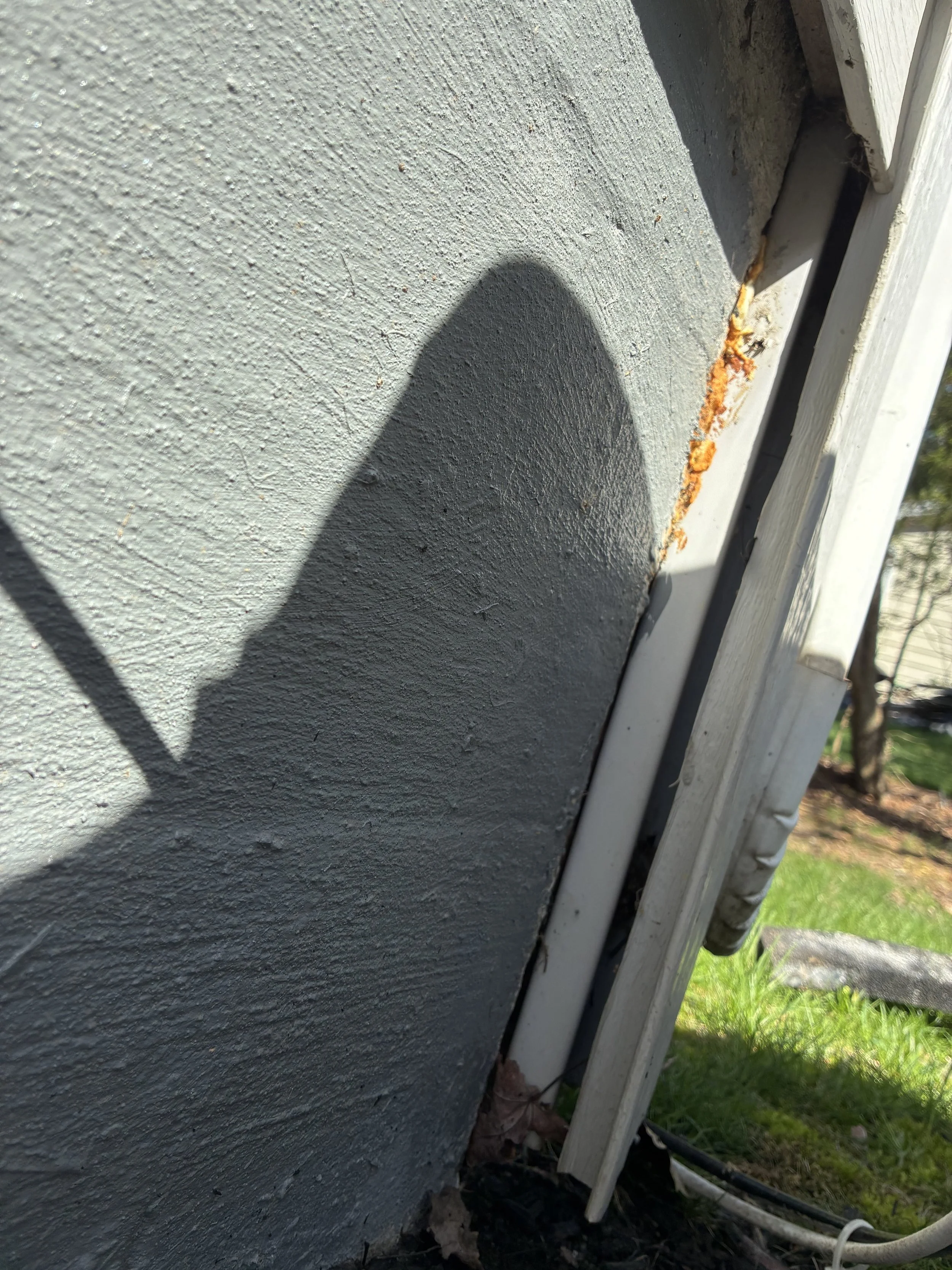 Close-up of a corner of a house with textured gray walls and a rusted metal pipe with peeling paint along the side. Grass and dirt are visible at the bottom corner.