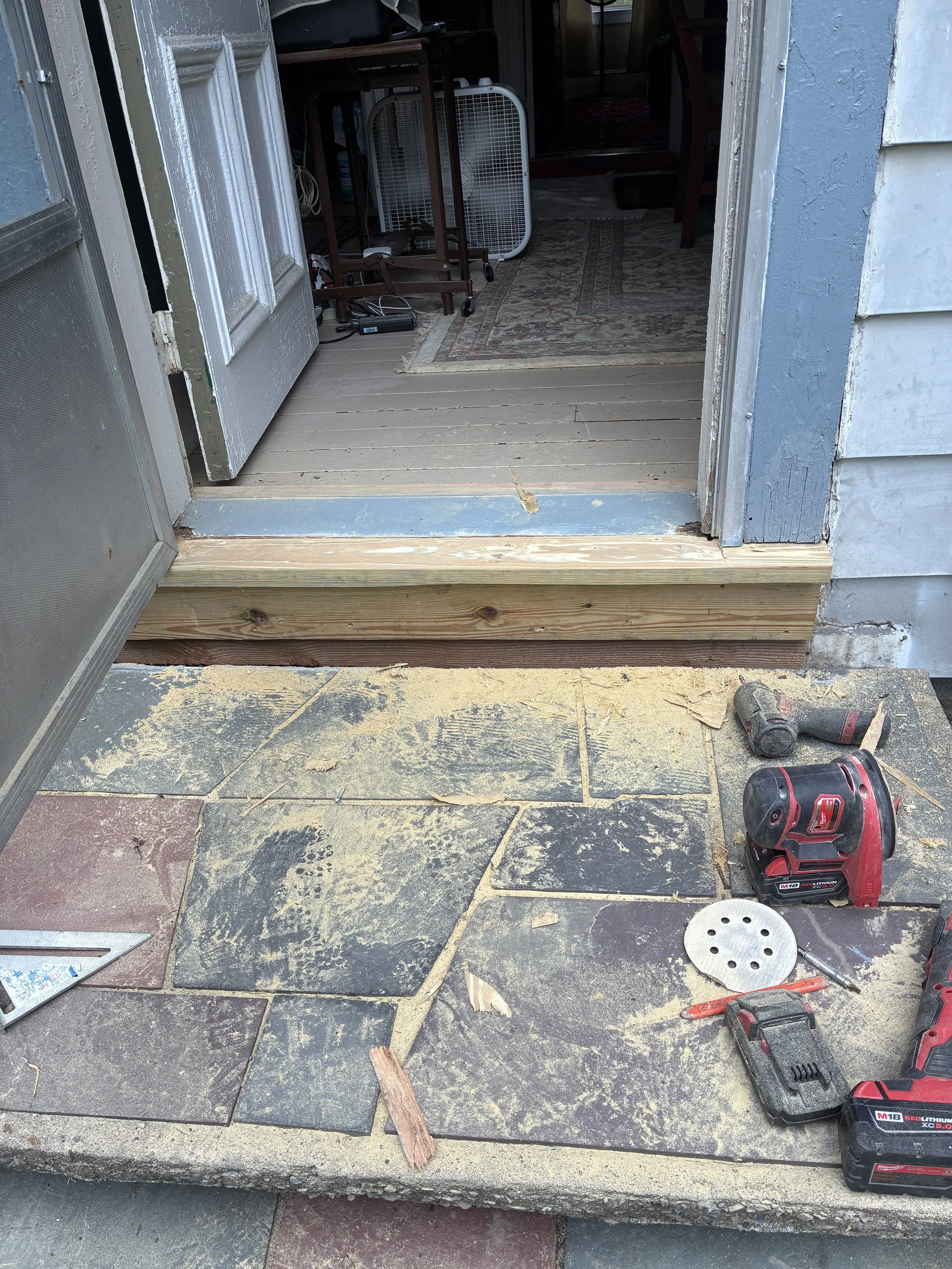 Exterior door opening to an indoor space with construction tools and materials, including a cordless drill, a round saw, wood pieces, and a cordless impact wrench, on a stone patio with construction debris.