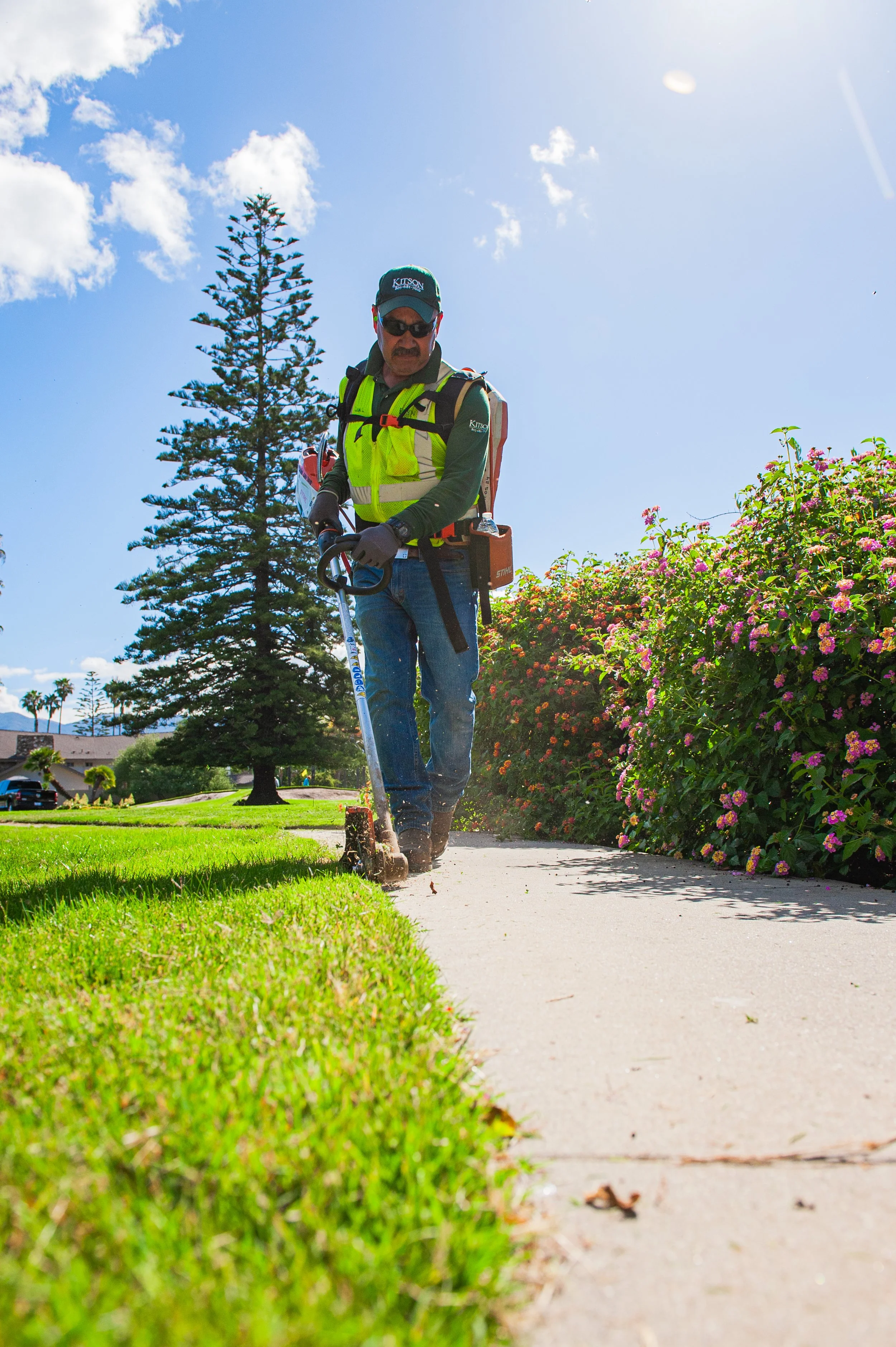 A man wearing sunglasses, a hat, gardening gloves, a bright yellow vest, and carrying a backpack, using a garden edger to trim the grass along a sidewalk on a sunny day. There are trees, flowering bushes, and houses in the background.