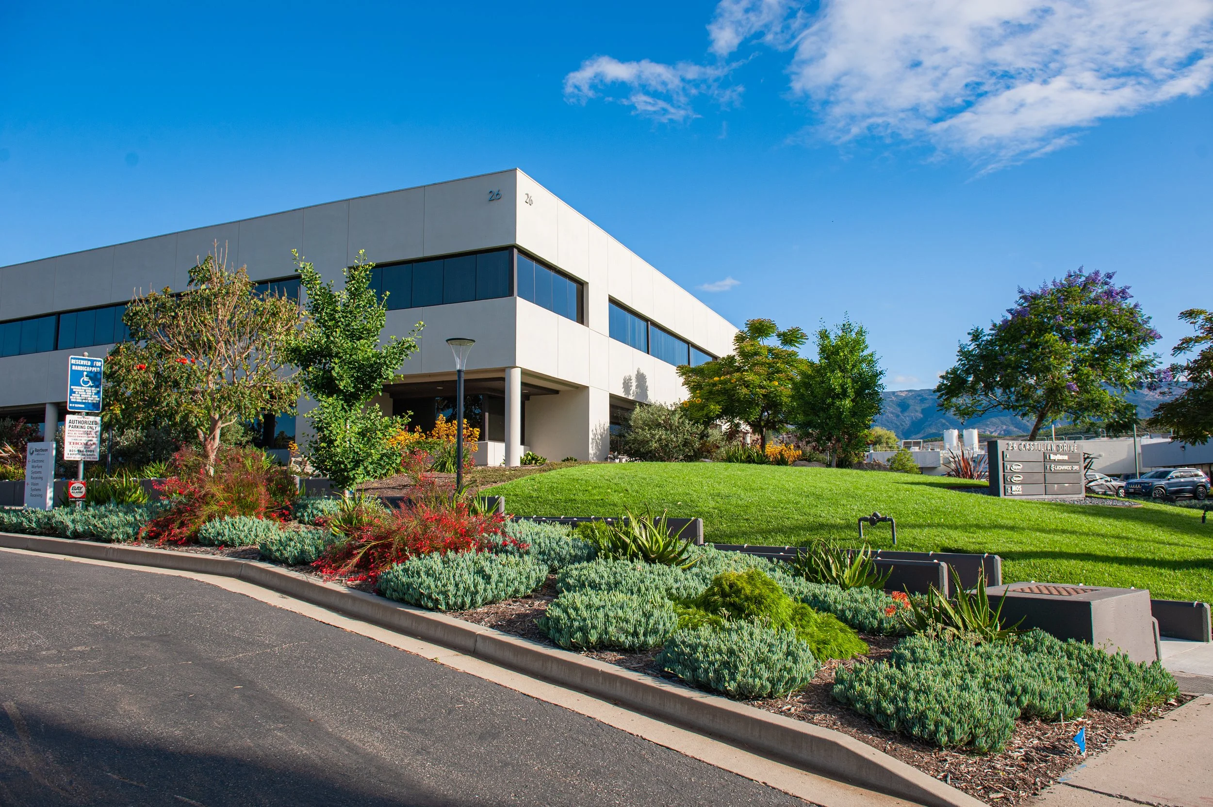 Modern white commercial building with large windows, surrounded by landscaped garden and trees under a blue sky with clouds.