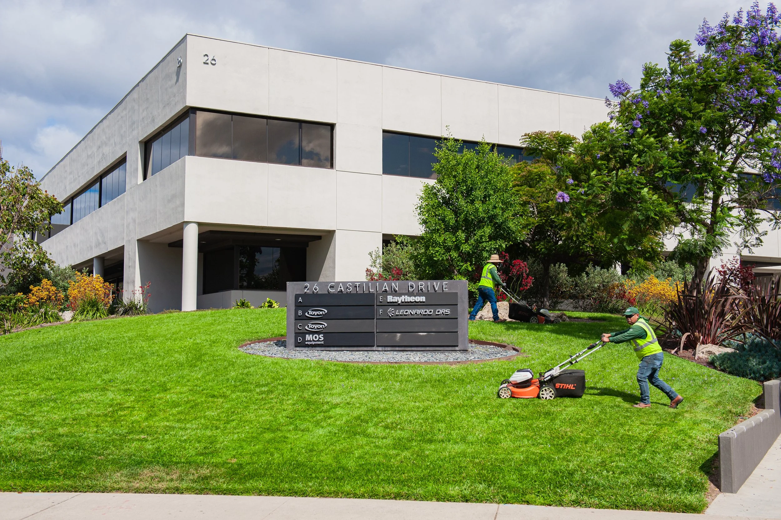 Modern white office building surrounded by well-maintained green grass and landscaped plants. Two workers in safety vests are mowing the lawn with a trimmer and a mower. A sign in front of the building displays company logos and names.