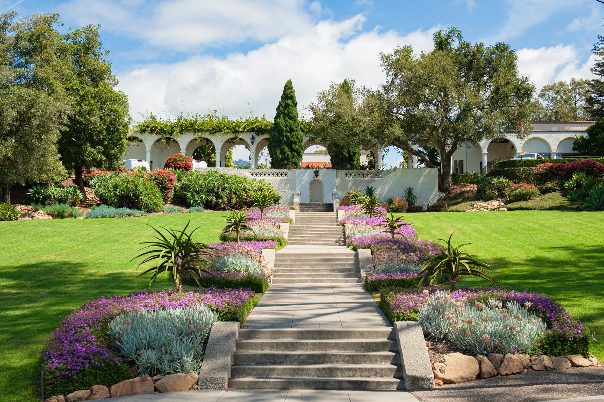 A set of concrete stairs leads up to a courtyard surrounded by lush greenery and colorful flowering bushes, with large trees and a white building with arches in the background under a partly cloudy blue sky.