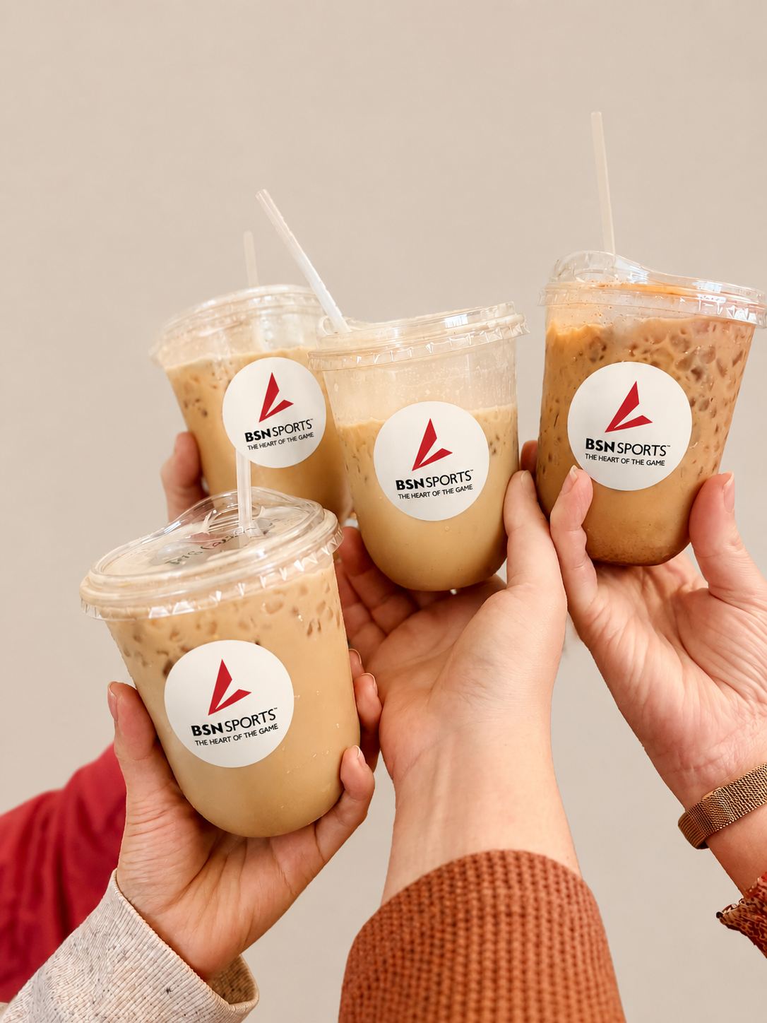 Four hands holding up iced coffee drinks with BSN Sports logos, celebrating together.