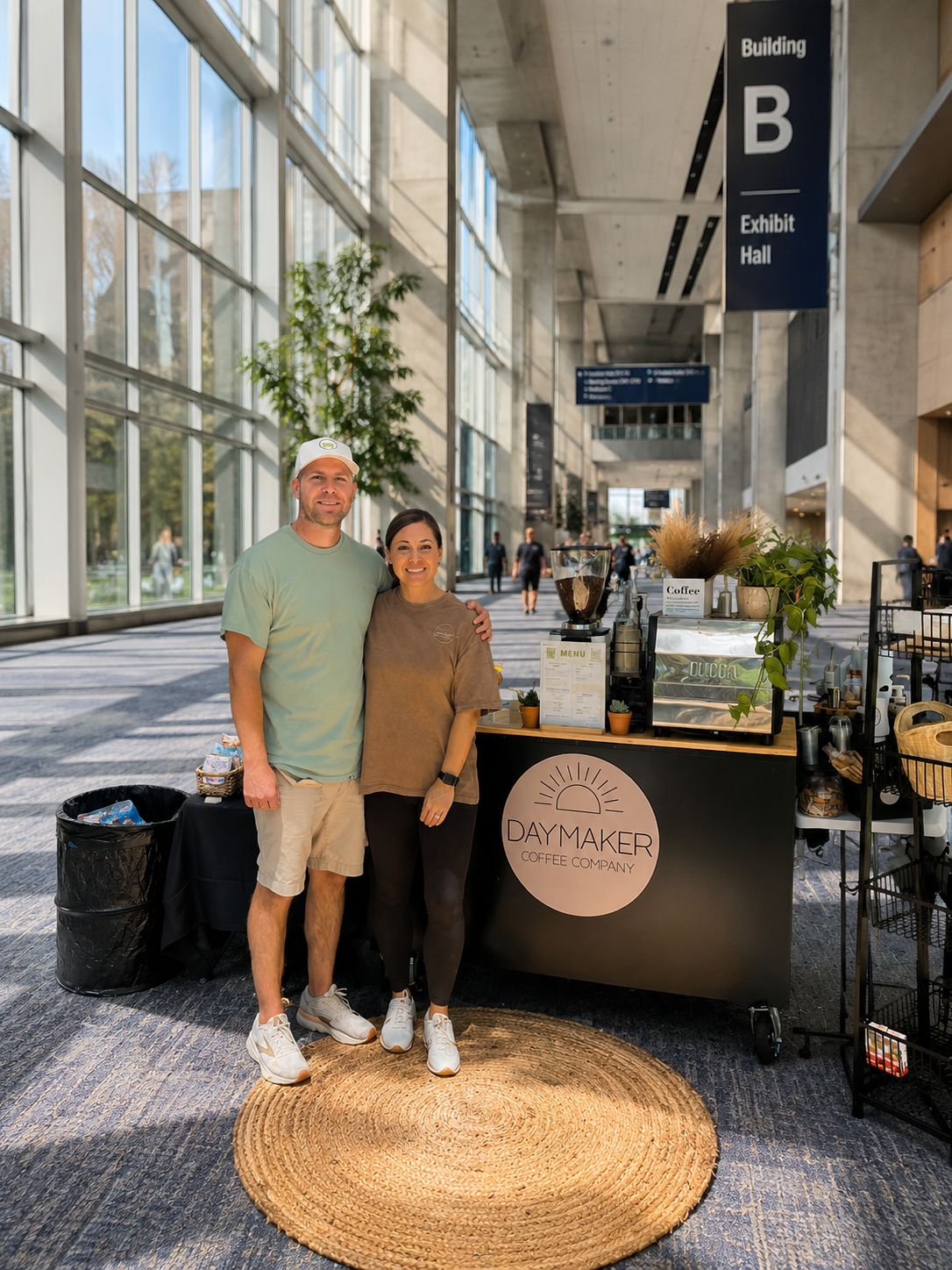 Owners of Daymaker Coffee Company standing beside their mobile coffee cart and espresso bar setup at a convention center event in Raleigh, NC.