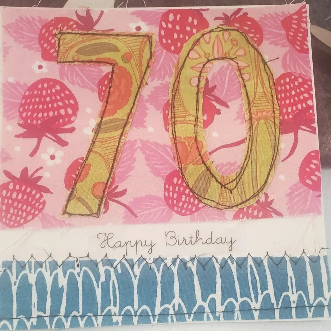 Handmade 70th birthday greeting card with stitched fabric appliqué numbers, colorful strawberry patterned paper, and layered cardstock accents.