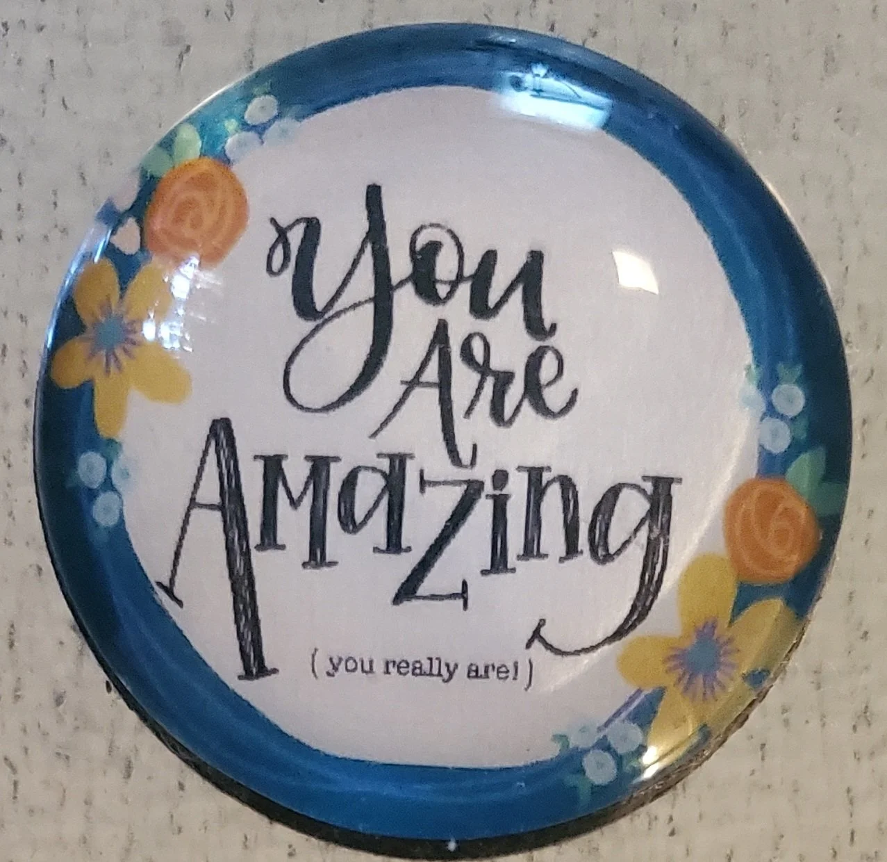 Handmade 2-inch round glass magnet featuring the words "You Are Amazing (you really are!)" surrounded by a bright blue floral border beneath a crystal-clear glass dome.