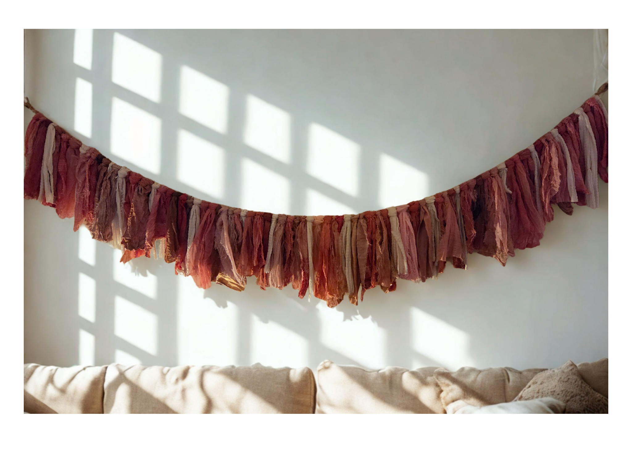 Handmade pink Chindi rug fabric garland in rose, mauve, blush and neutral tones hanging on a sunlit wall above a cream sofa.