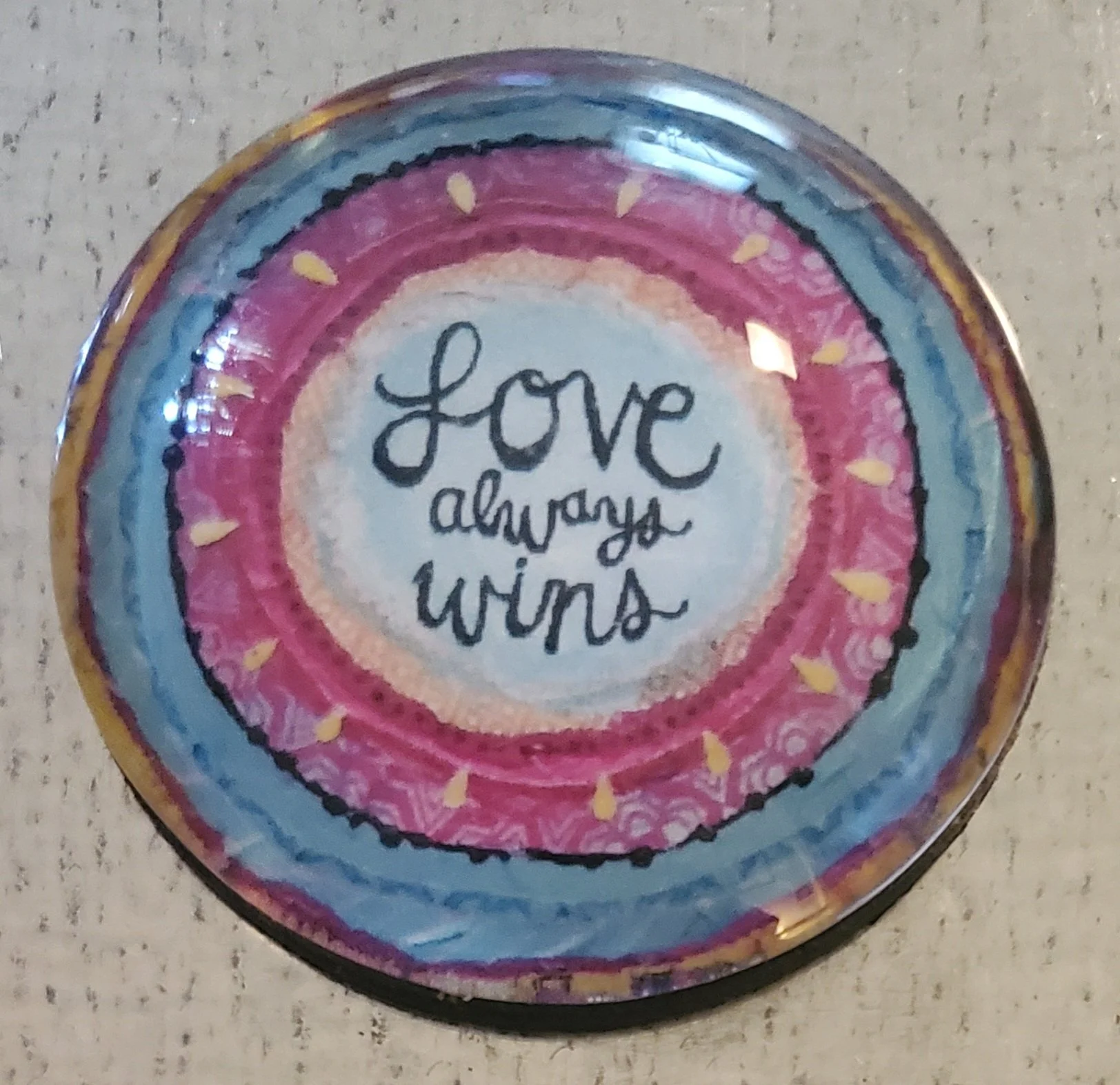 Handmade 2-inch round glass magnet featuring the phrase "Love Always Wins" surrounded by colorful watercolor-inspired concentric circles beneath a crystal-clear glass dome.