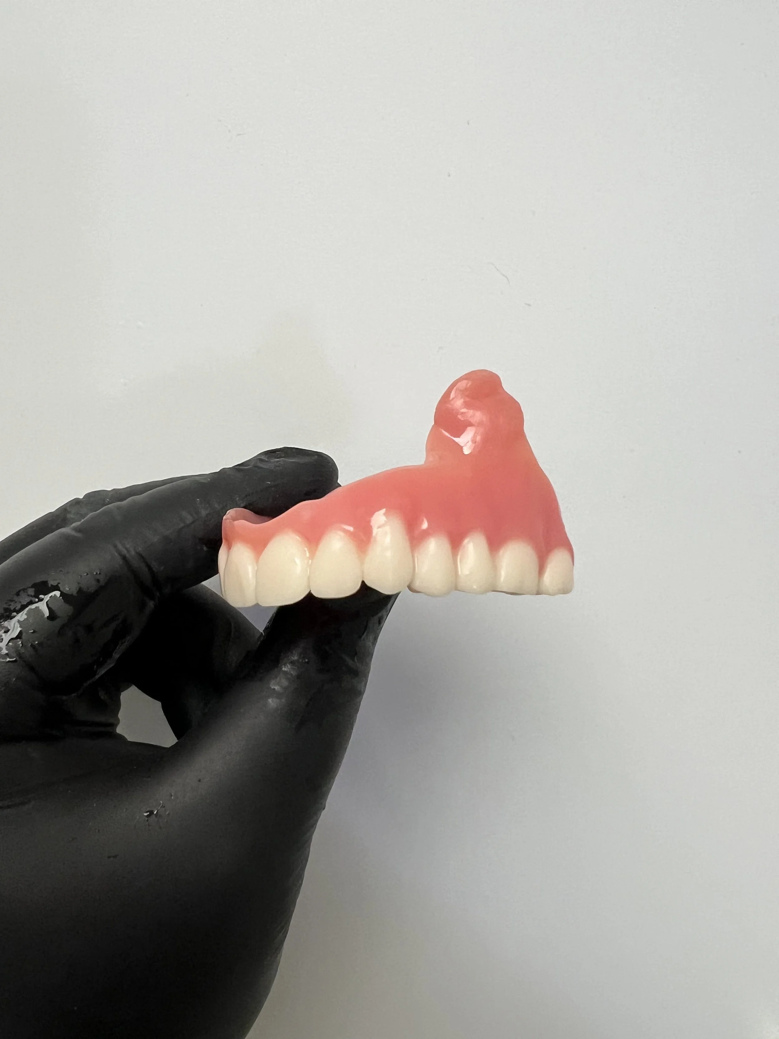 A hand wearing a black glove holding a partial dental prosthesis with artificial teeth and pink gum material against a light gray background.
