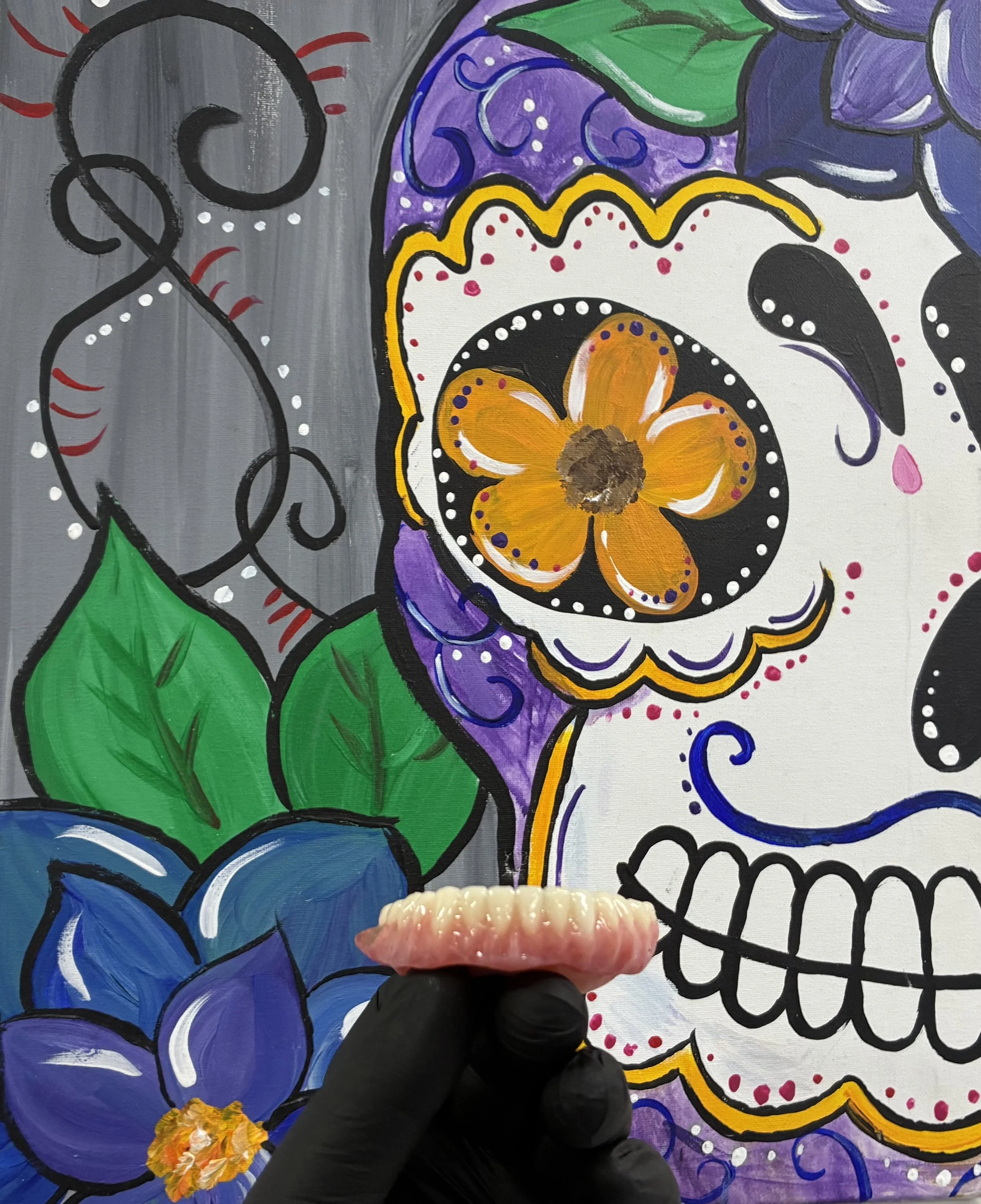A colorful painting of a sugar skull with flower and leaf designs, featuring purple, orange, green, black, purple, and white colors, with a person's hand with a ring holding a small flower in front of the artwork.