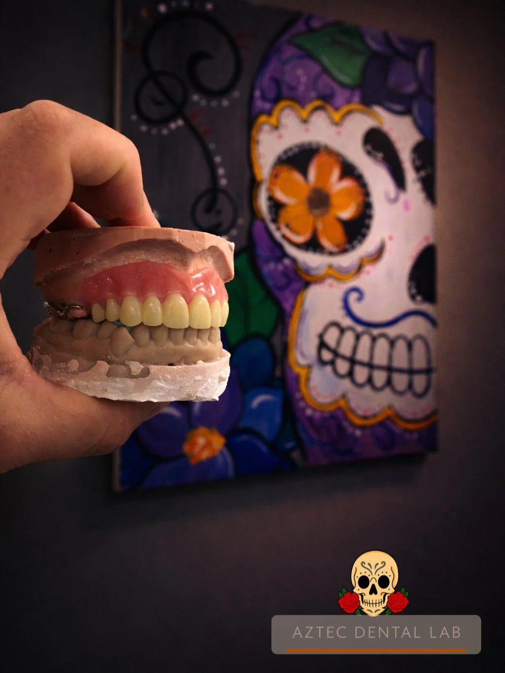A hand holding a dental prosthetic with pink gums and yellow teeth, in front of a colorful skull artwork, with the text 'AZTEC DENTAL LAB' and a skull with roses logo at the bottom.