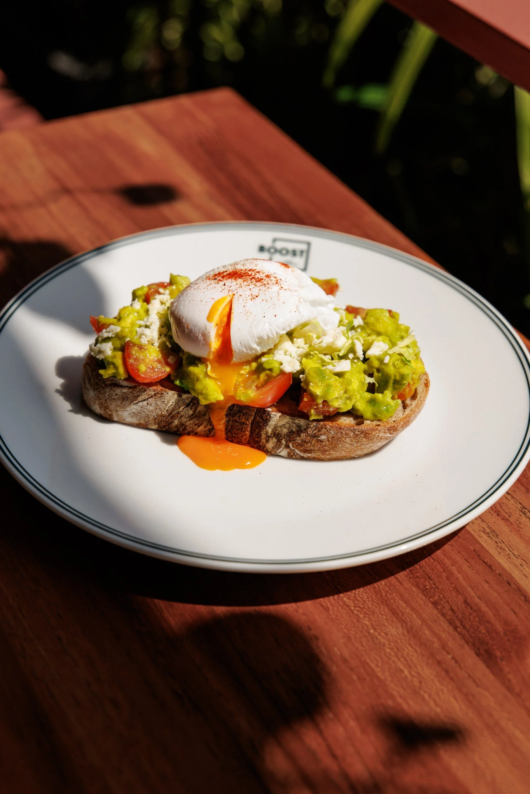Open-faced avocado toast with cherry tomatoes, crumbled cheese, and a poached egg on top, served on a white plate.