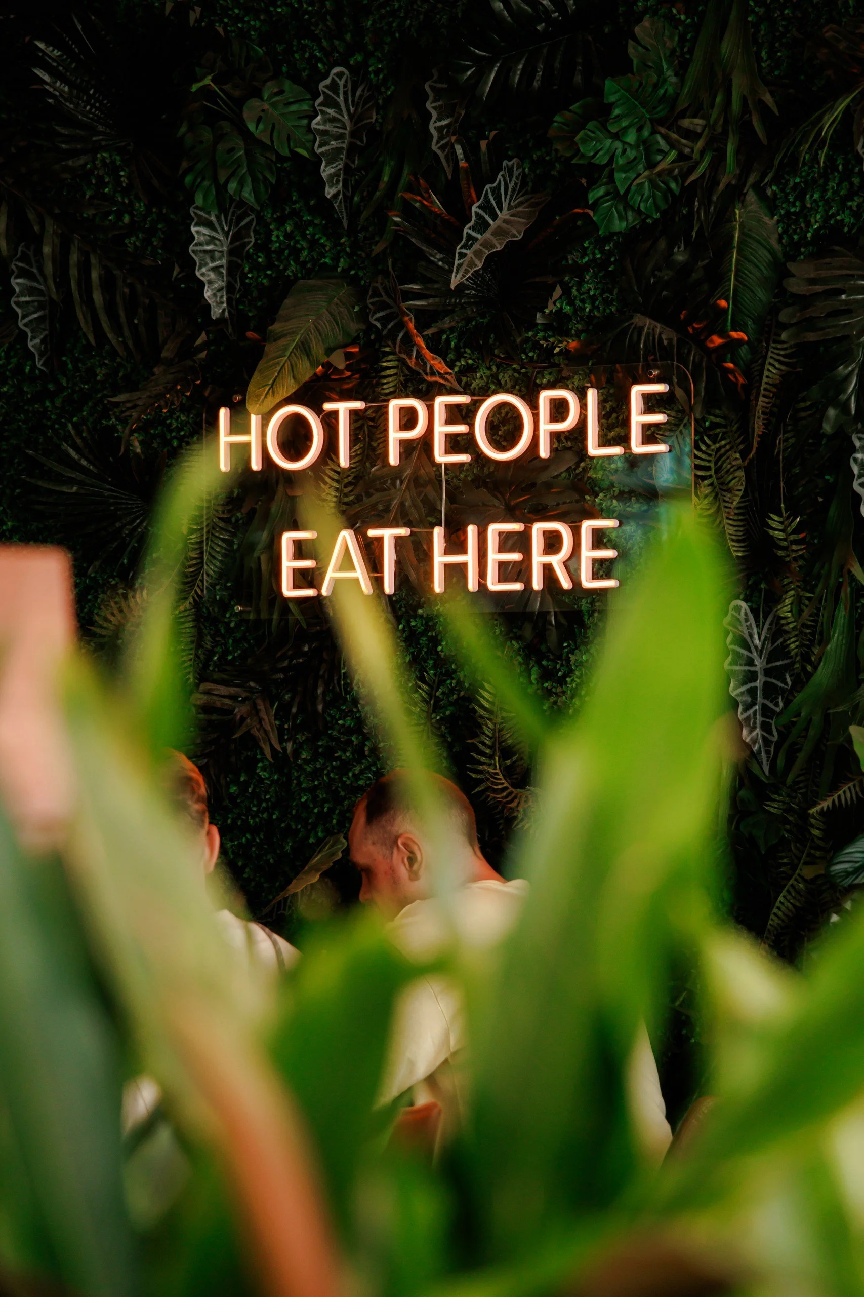A neon sign that reads 'HOT PEOPLE EAT HERE' against a background of tropical plants, with a few people visible in the lower part of the image.