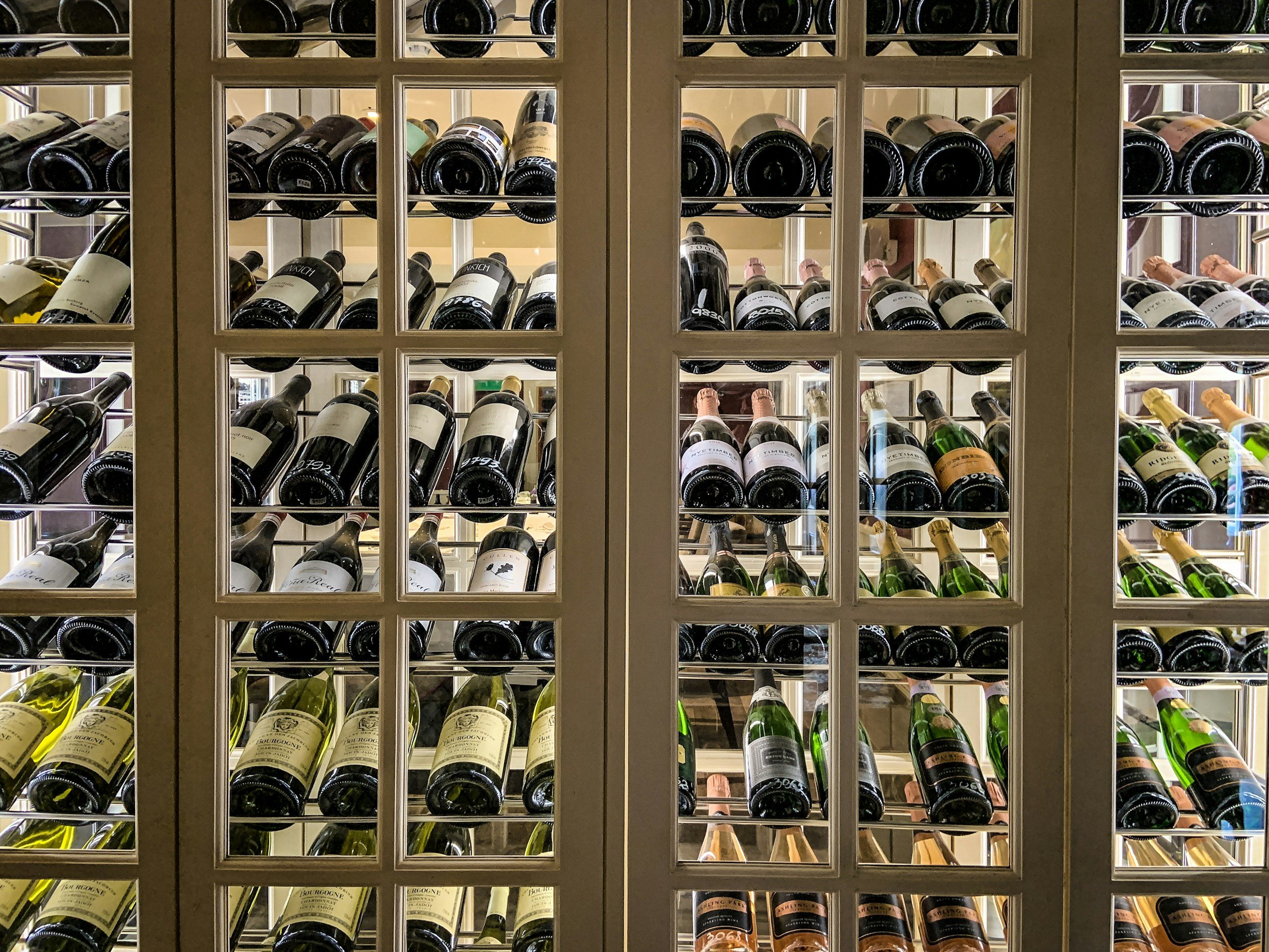 Wine bottles stored horizontally in a wine cellar with a glass-fronted wine rack in the foreground.