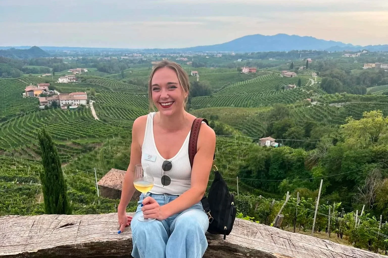 A woman with a glass of white wine sitting on a wooden bench in front of rolling vineyards and hills with a cloudy sky in the background.