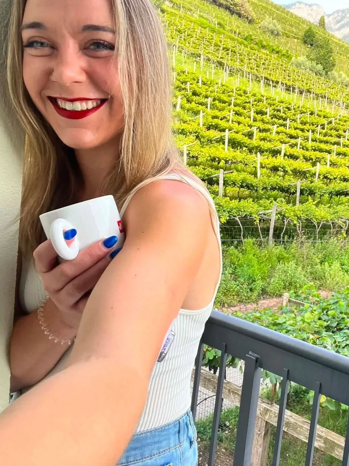 A woman smiling and holding a white mug with red and blue details on a balcony, with a lush green vineyard or hillside in the background.