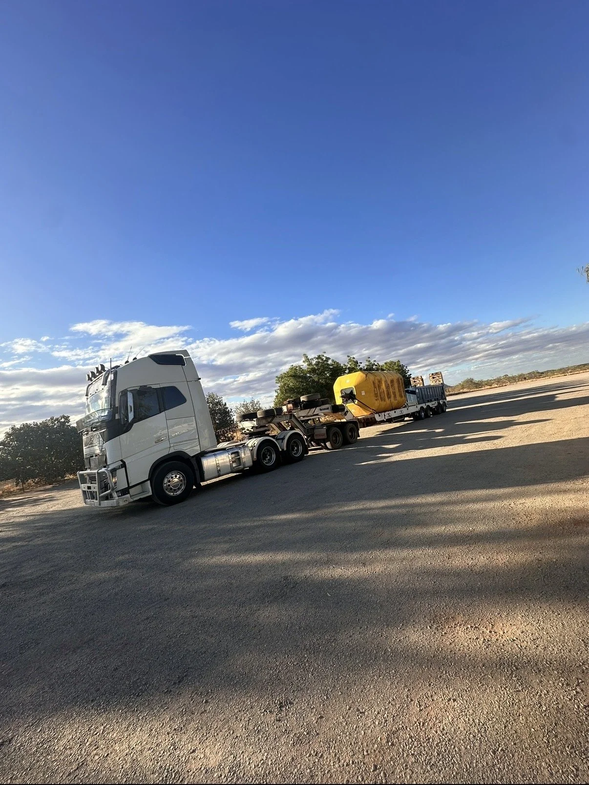 A white semi-truck parked on the side of a paved road, with a trailer carrying large yellow and black containers, under a partly cloudy sky with trees in the background.