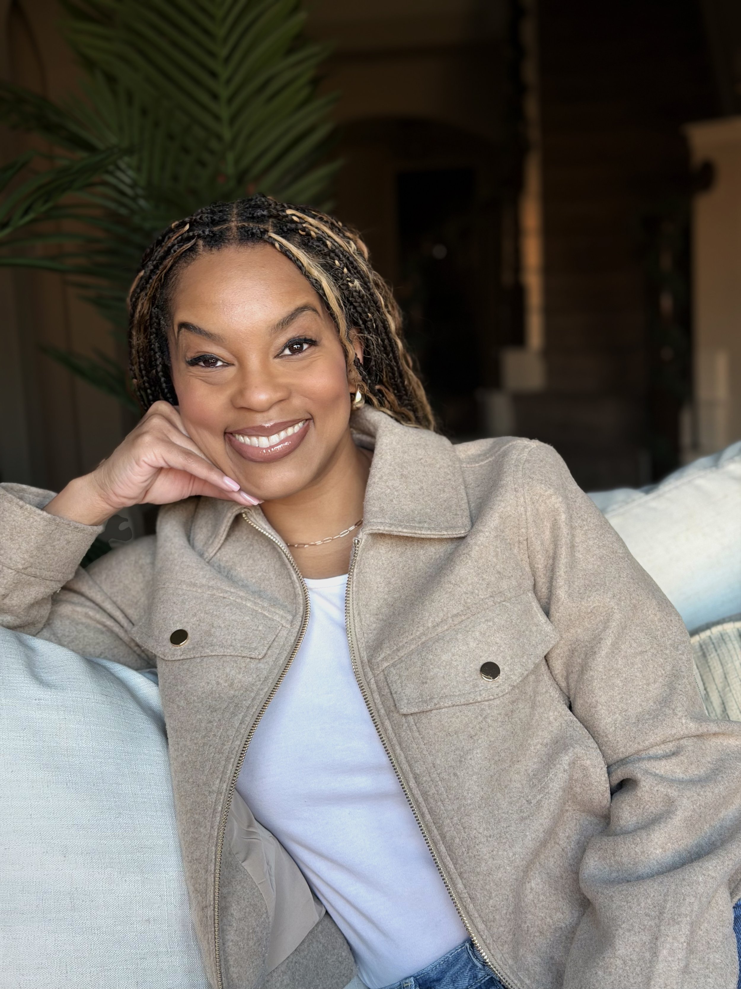 A woman with braided hair smiling and resting her chin on her right hand, sitting on a light blue couch, with a large green plant in the background.